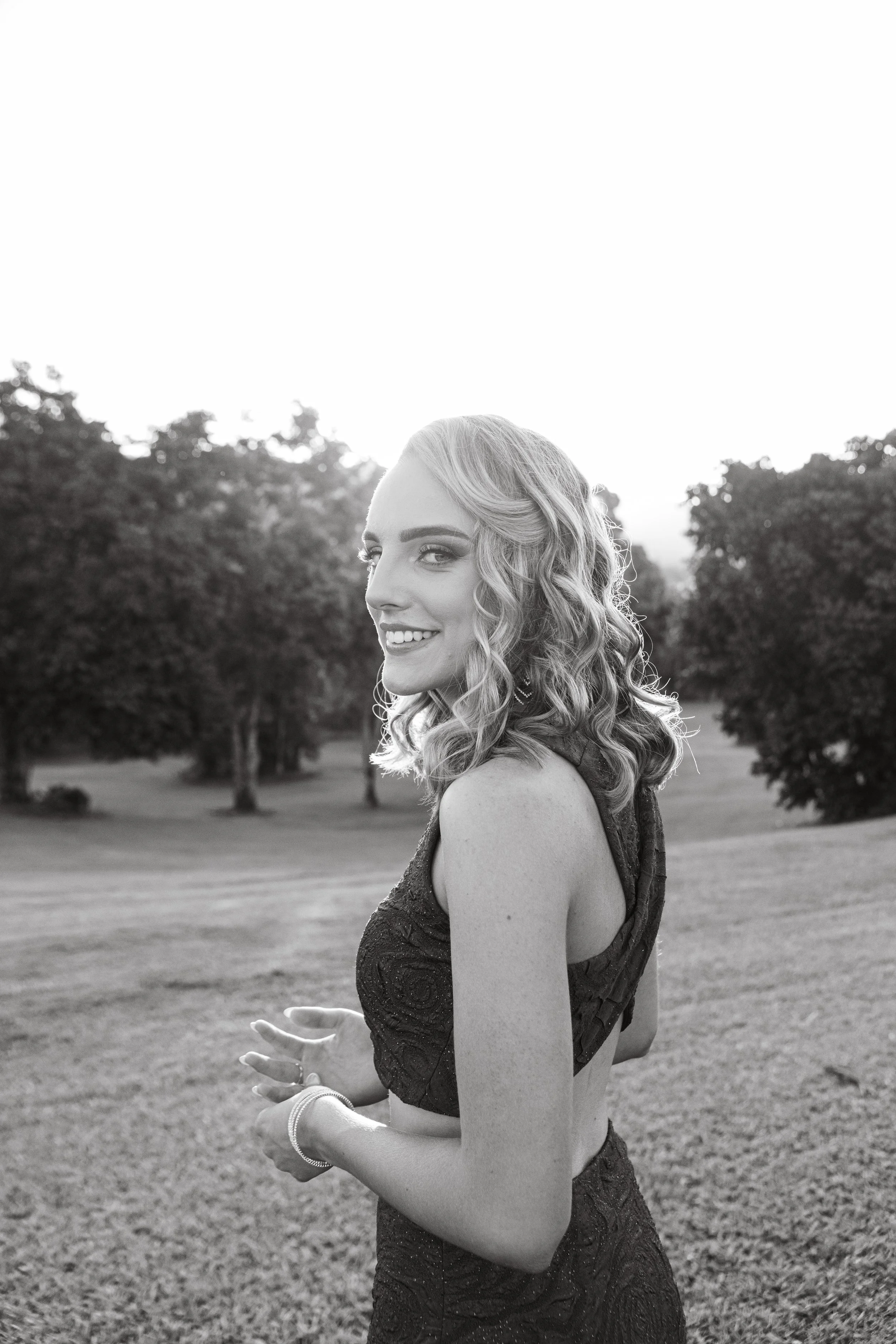 A young woman with wavy hair smiling outdoors in a black dress, standing on a grassy area with trees in the background, in black and white.