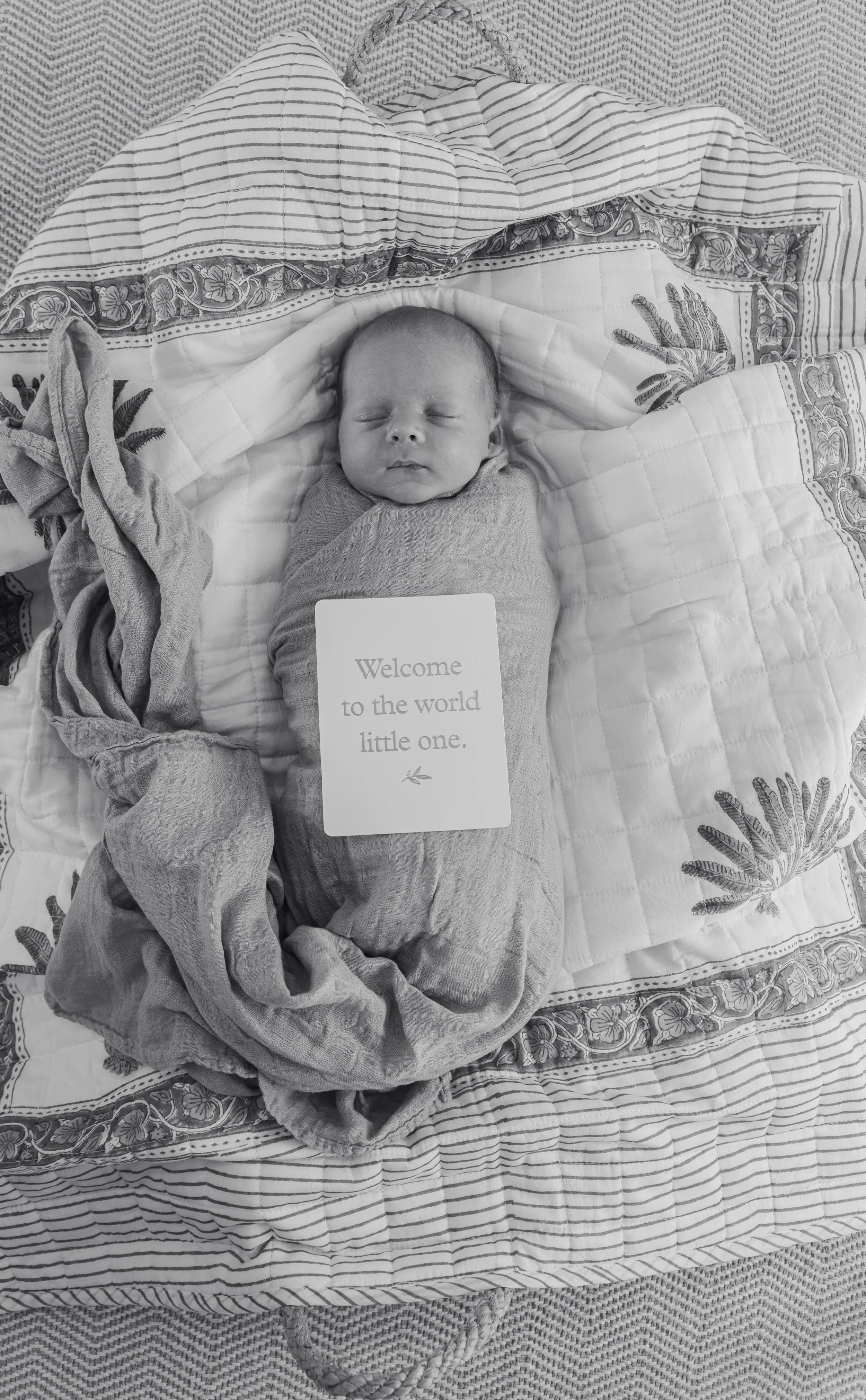 A newborn baby sleeping on a quilted blanket, wrapped in a cloth, with a card that reads "Welcome to the world little one."