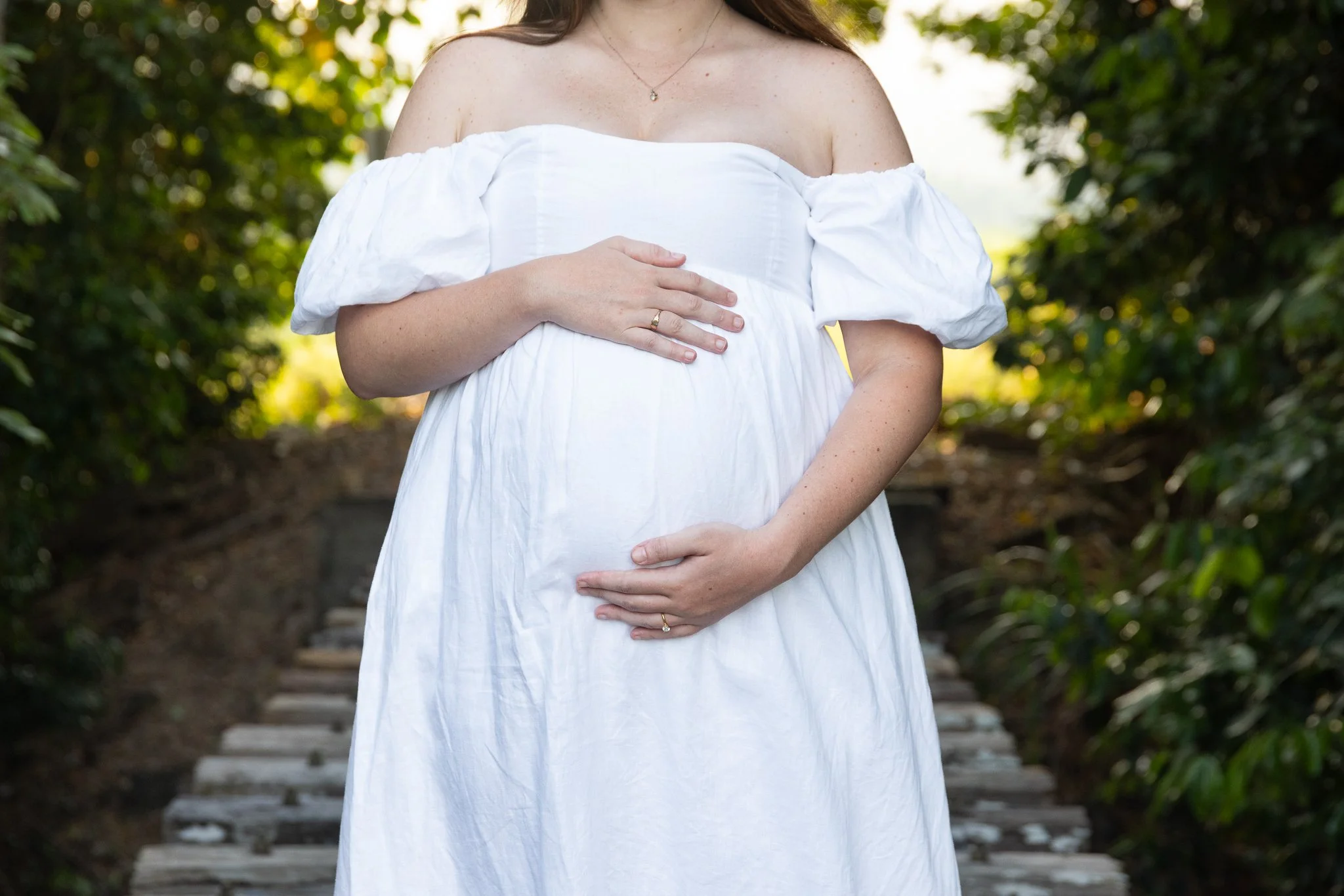 A pregnant woman wearing a white off-the-shoulder dress stands outdoors on a wooden walkway, with hands on her belly, surrounded by green foliage.