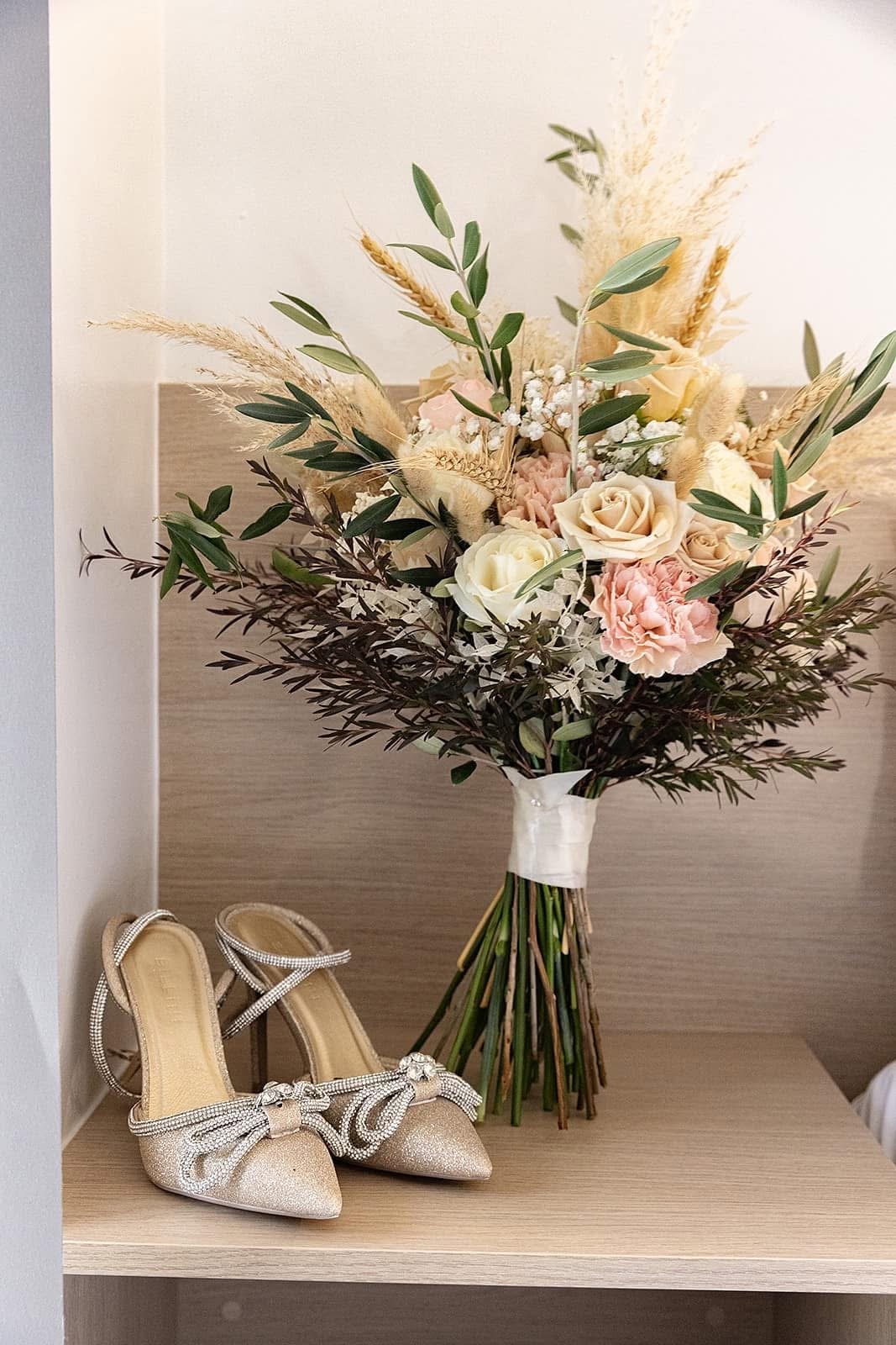 A bouquet of pink, white, and beige flowers with greenery, placed on a shelf next to a pair of glittery beige high heels with bows.