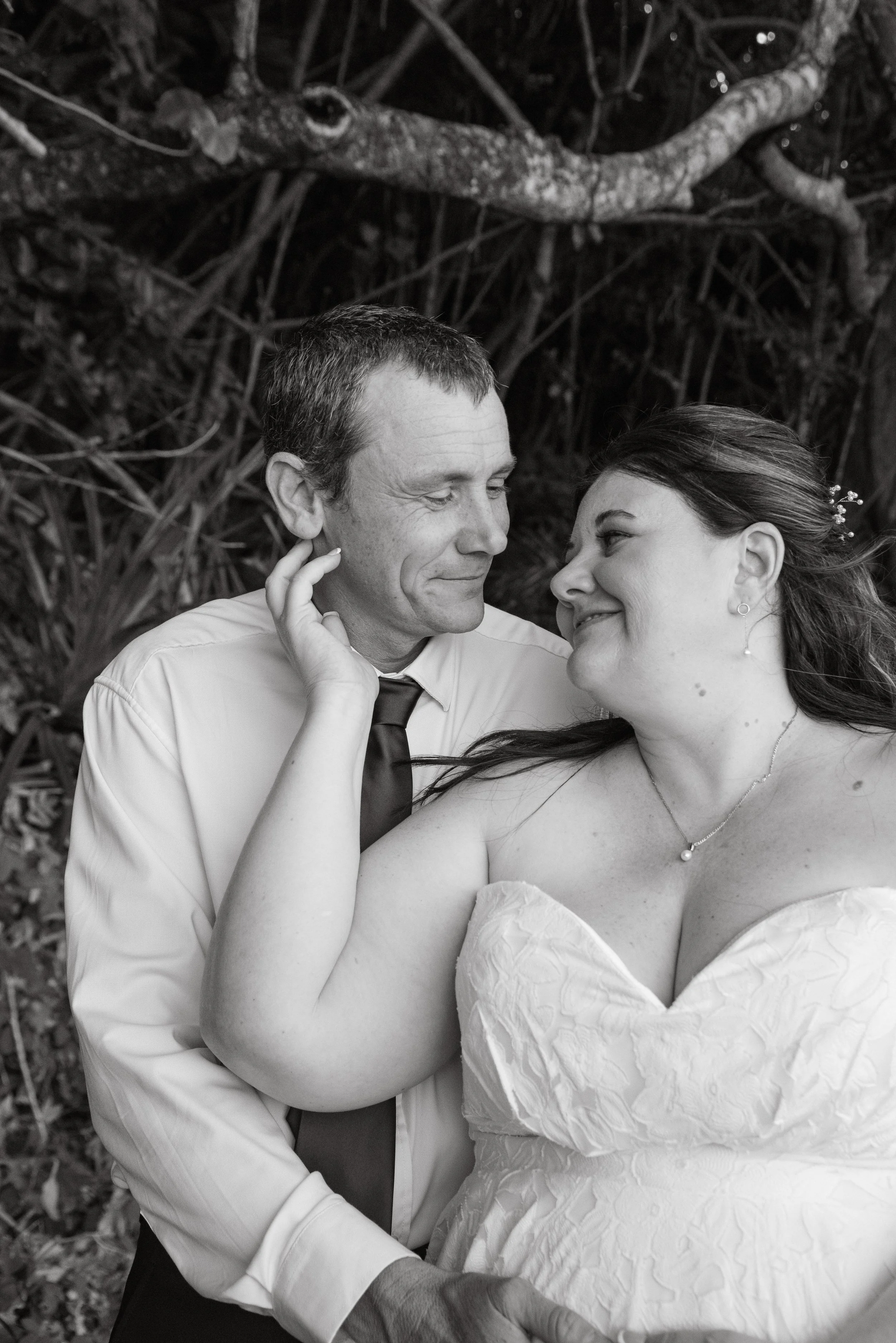 A black-and-white photo of a bride and groom gazing at each other, with the bride touching the groom's face, in an outdoor setting with trees in the background.