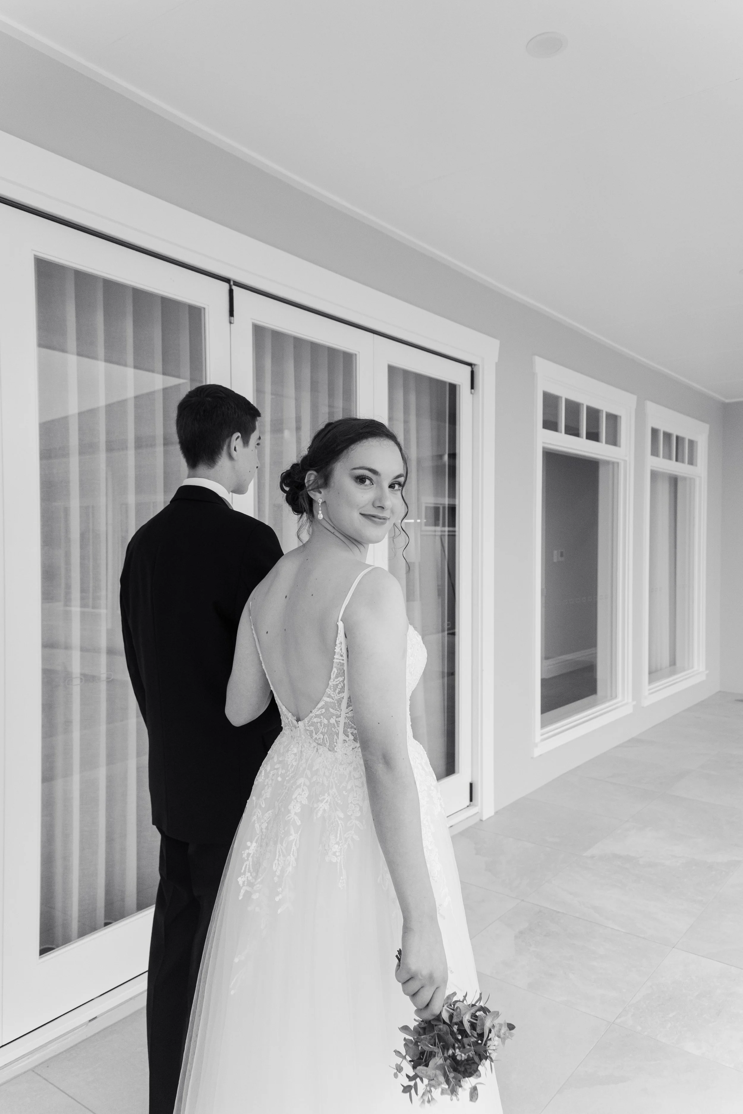 Black and white photo of a bride in a wedding dress holding a bouquet standing in front of a groom in a suit, inside a room with large windows.