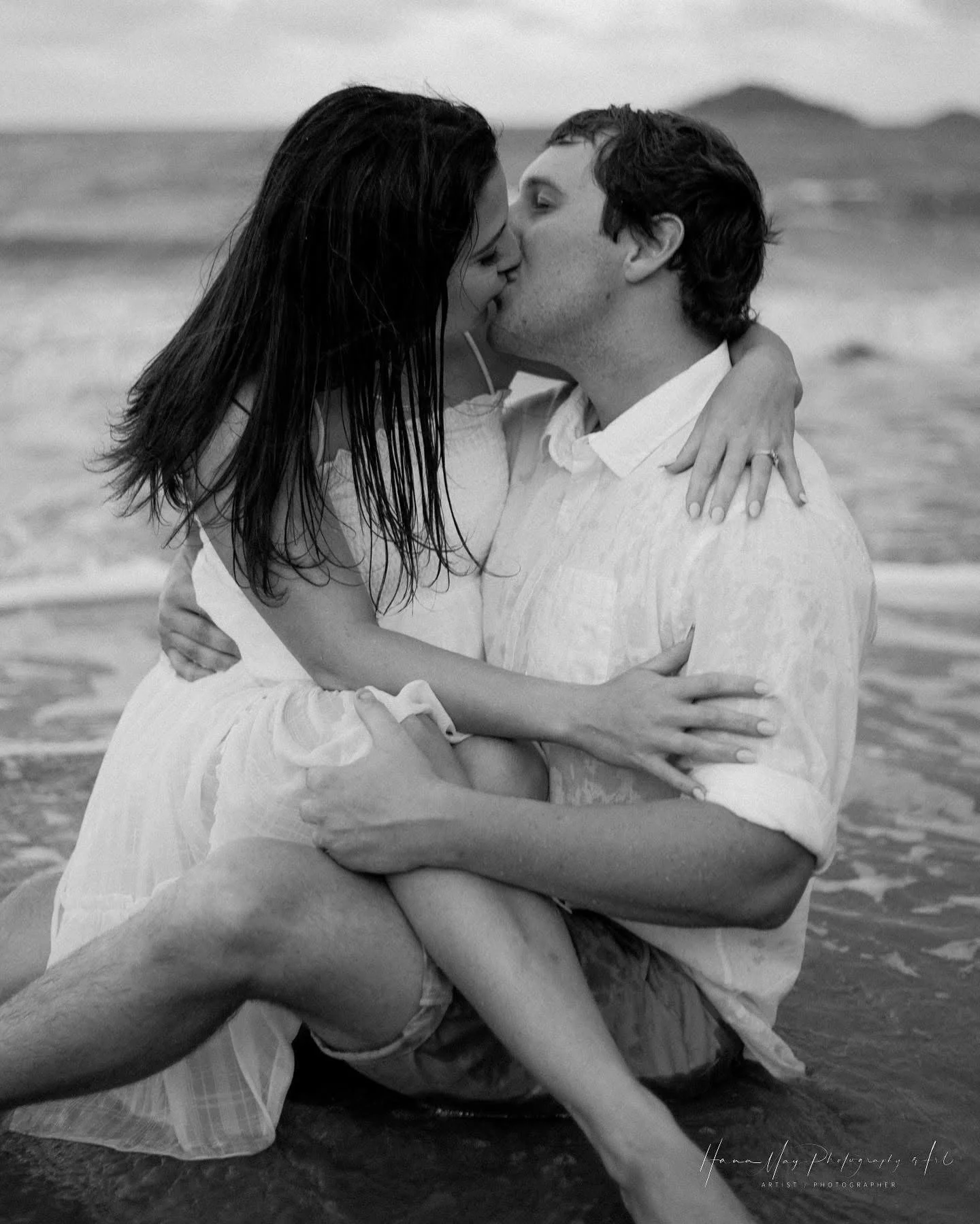 A black and white photo of a couple kissing on a beach, with the woman sitting on the man's lap and both embracing each other.