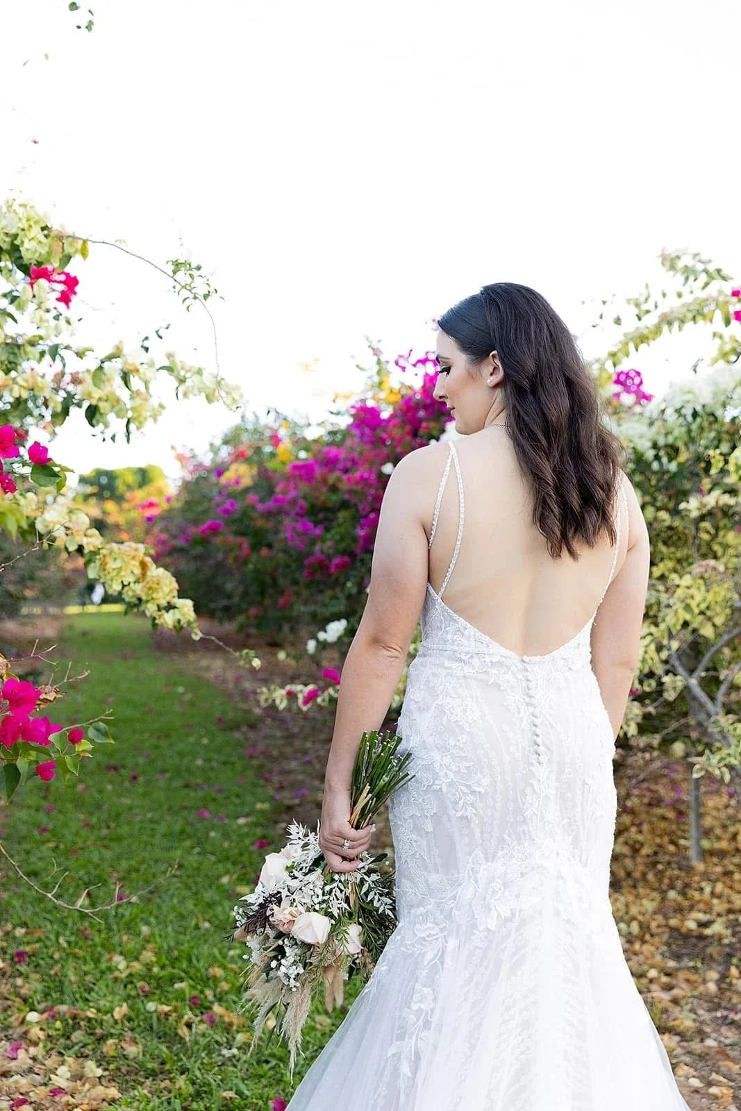 A bride in a white lace wedding dress with thin straps holding a bouquet of flowers, standing in a vibrant garden with blooming pink, yellow, and white flowers.