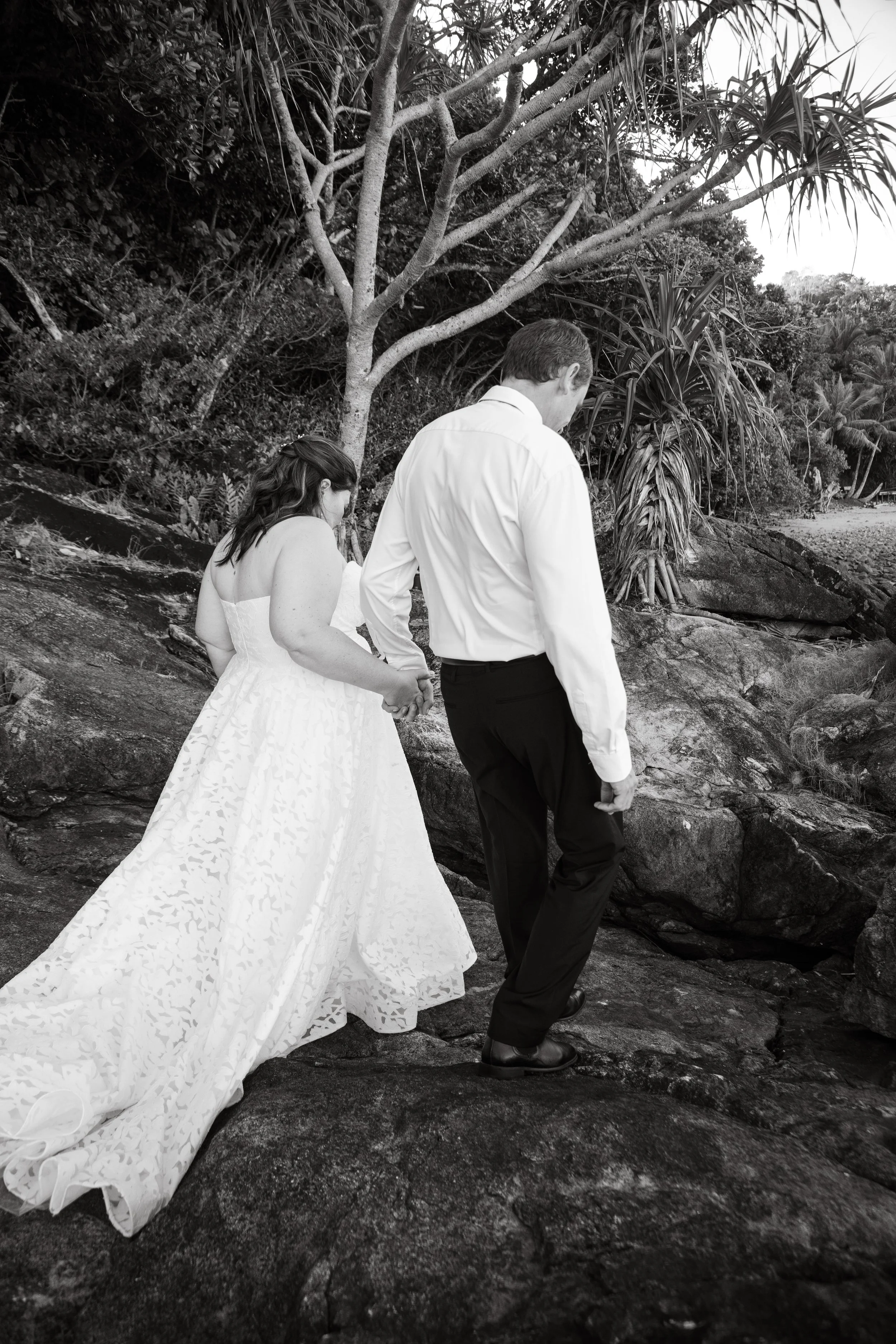 A couple, the woman in a white wedding dress and the man in a white shirt and dark trousers, holding hands while walking on rocky terrain near the water with trees and vegetation in the background.
