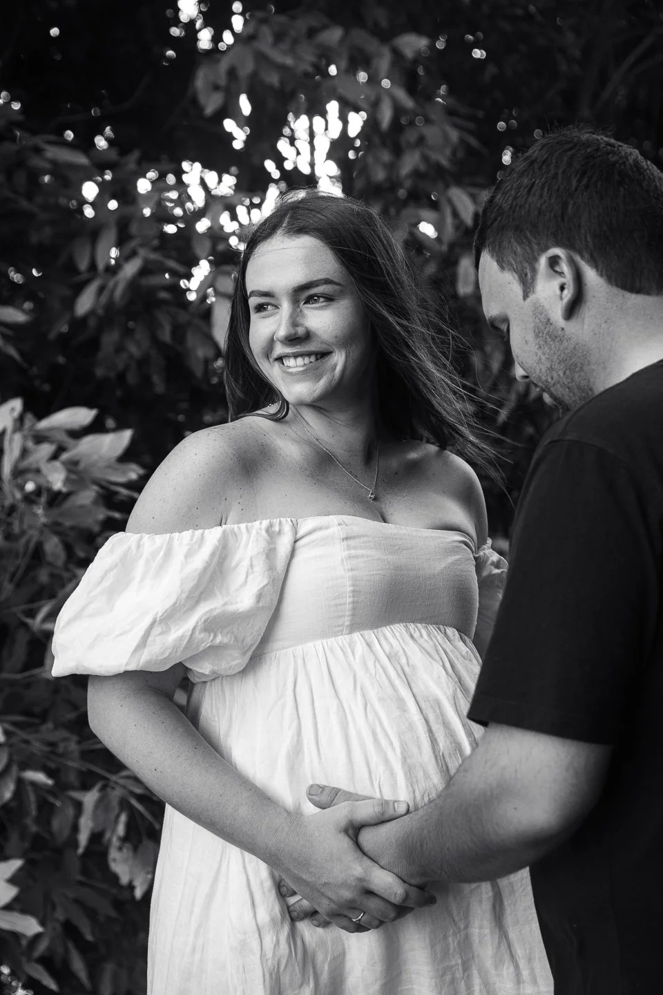 A happy pregnant woman smiling and looking away, holding hands with her partner outdoors with leafy background, black and white photo.