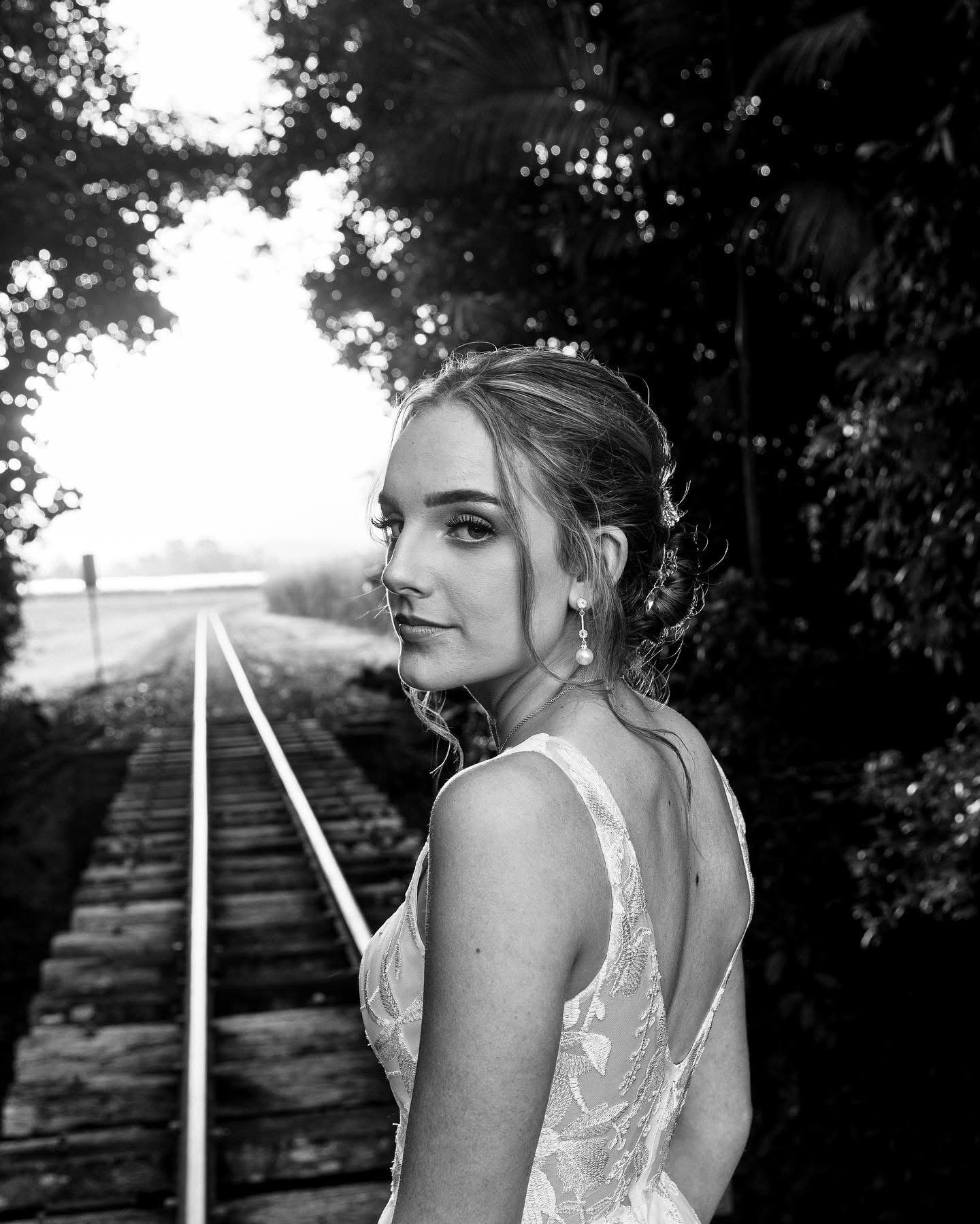 A black and white photo of a young woman in a lace dress, standing on train tracks among trees, looking over her shoulder with a soft smile.