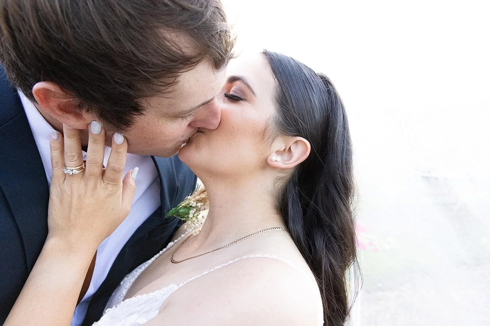 A bride and groom kissing during their wedding, with the bride wearing a white dress and jewelry, and the groom in a dark suit.