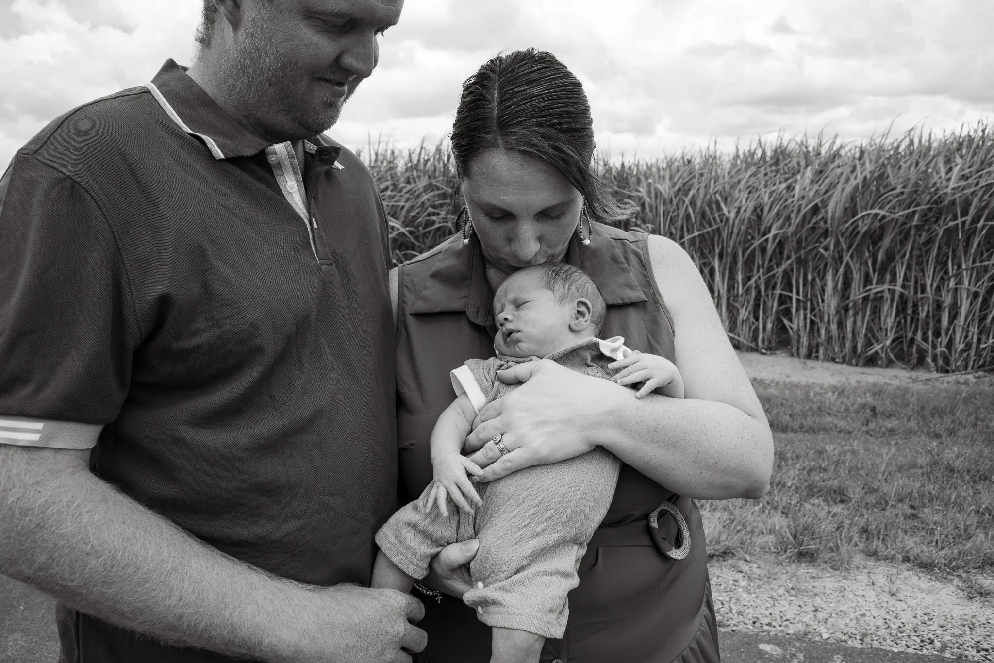 A woman holding a sleeping baby, with two adults looking on, outdoors near a field of tall grass or corn.