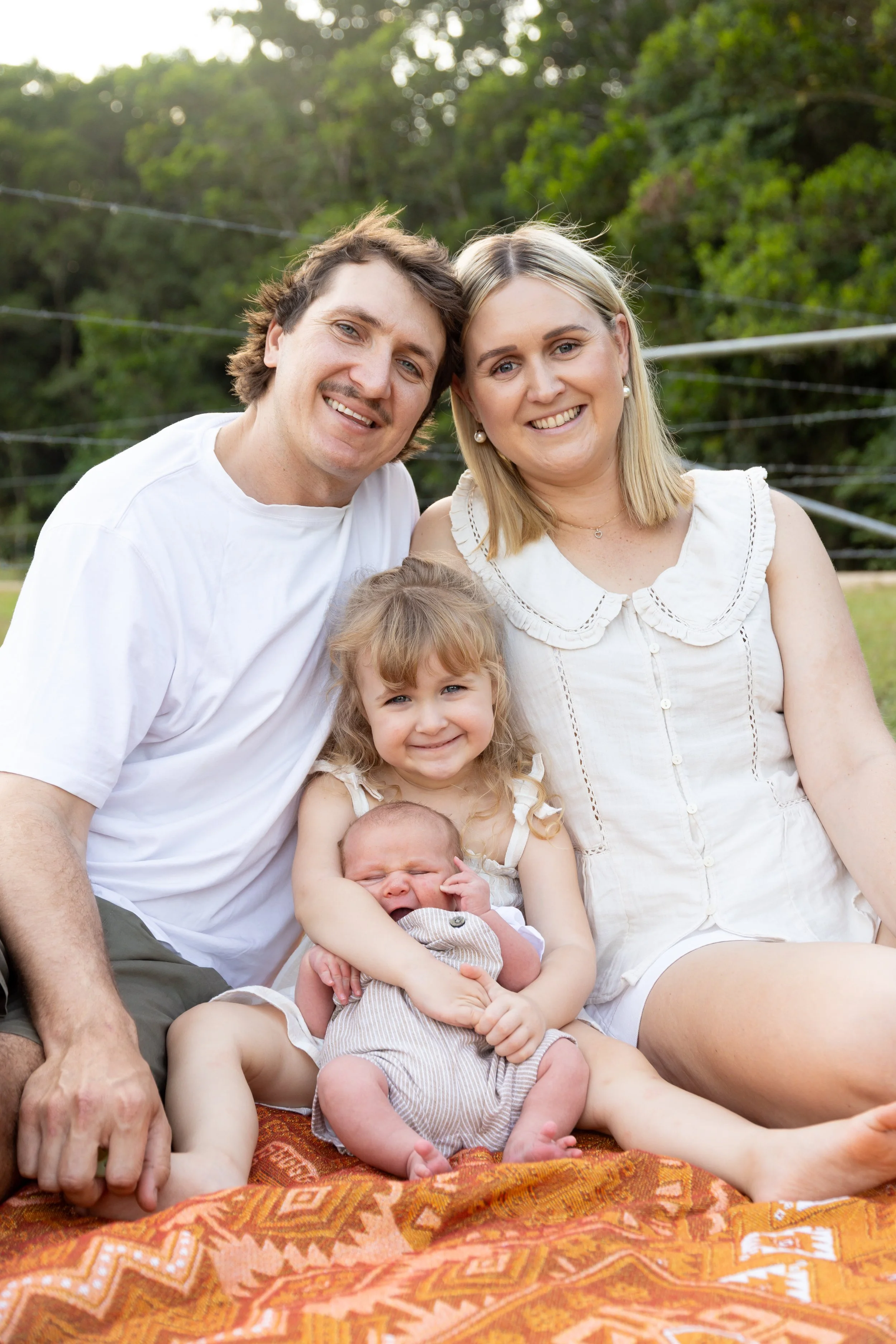A family of five sitting outdoors on a blanket, smiling at the camera, with trees and greenery in the background. The family includes a man, woman, two young girls, and a newborn baby.