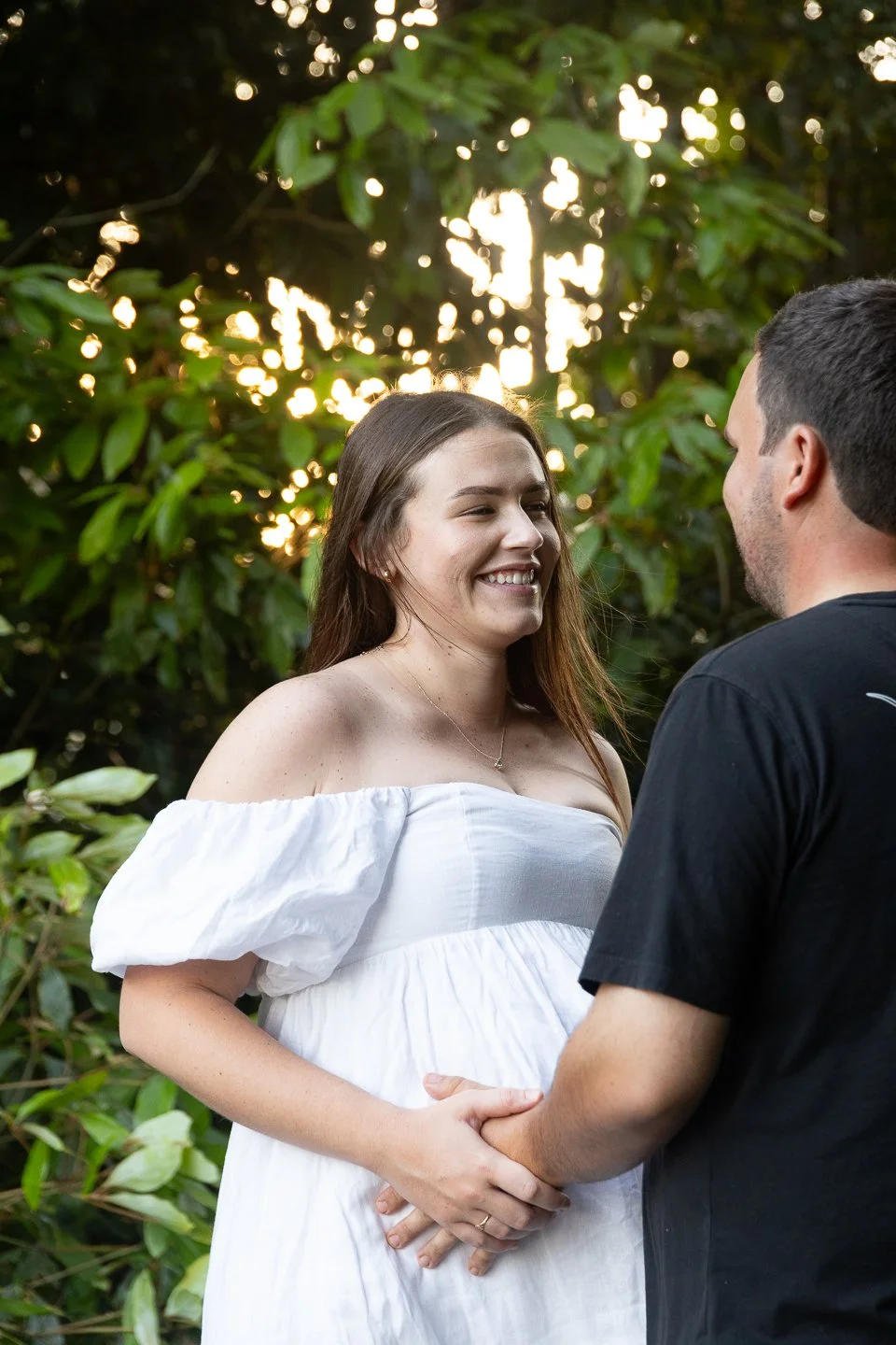 A woman and man holding hands and gazing at each other in a green outdoor setting with sunlight filtering through trees.