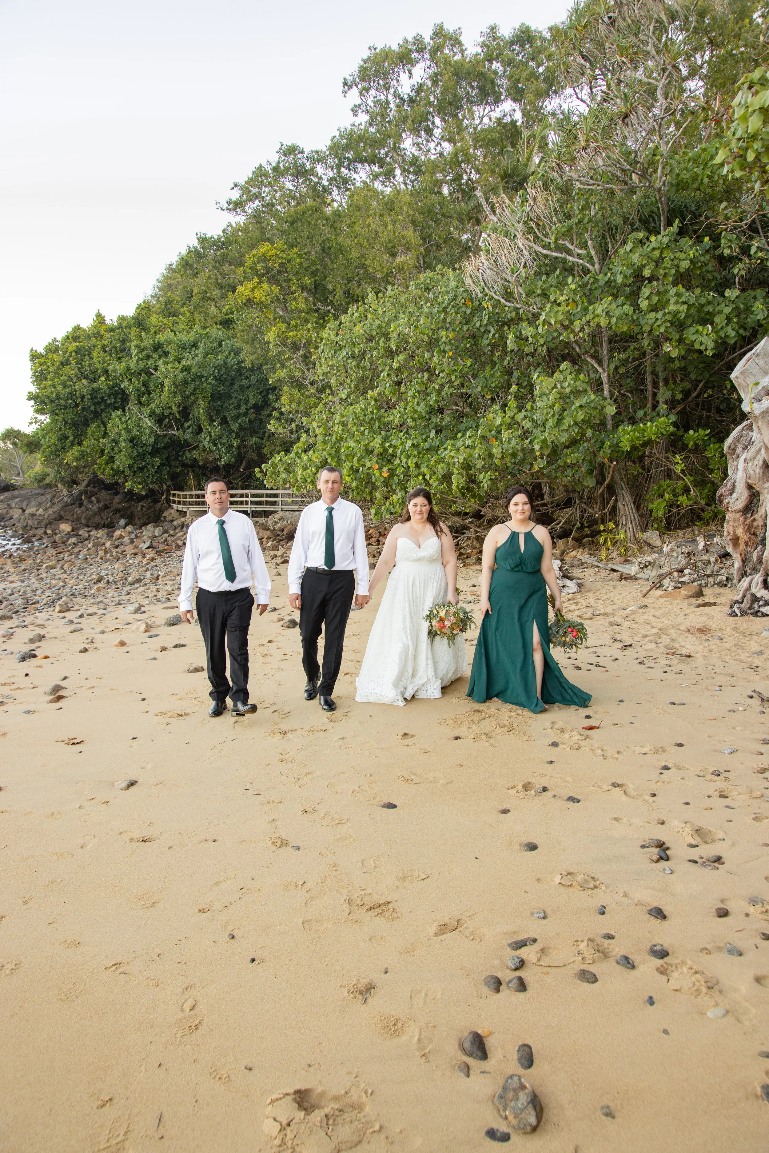 A wedding party walking on a sandy beach with lush green trees in the background.