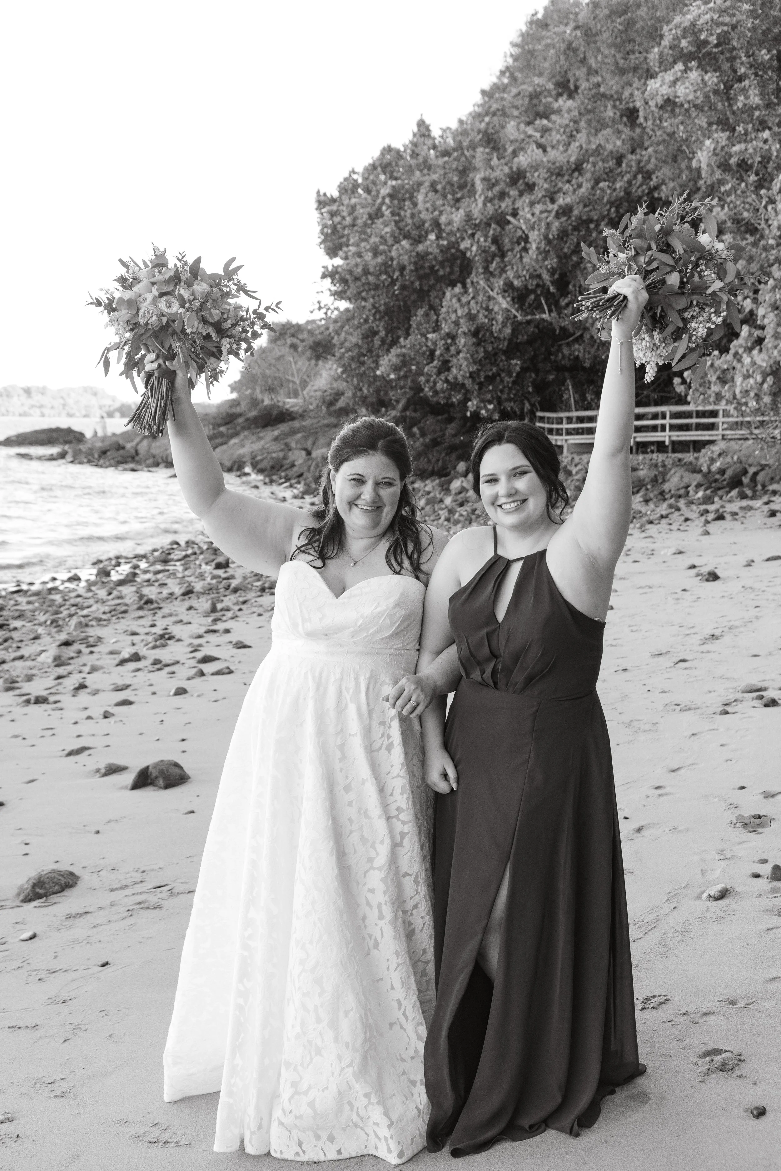 Two women in dresses celebrating on a beach, holding bouquets with arms raised, smiling, with trees and rocks in the background.