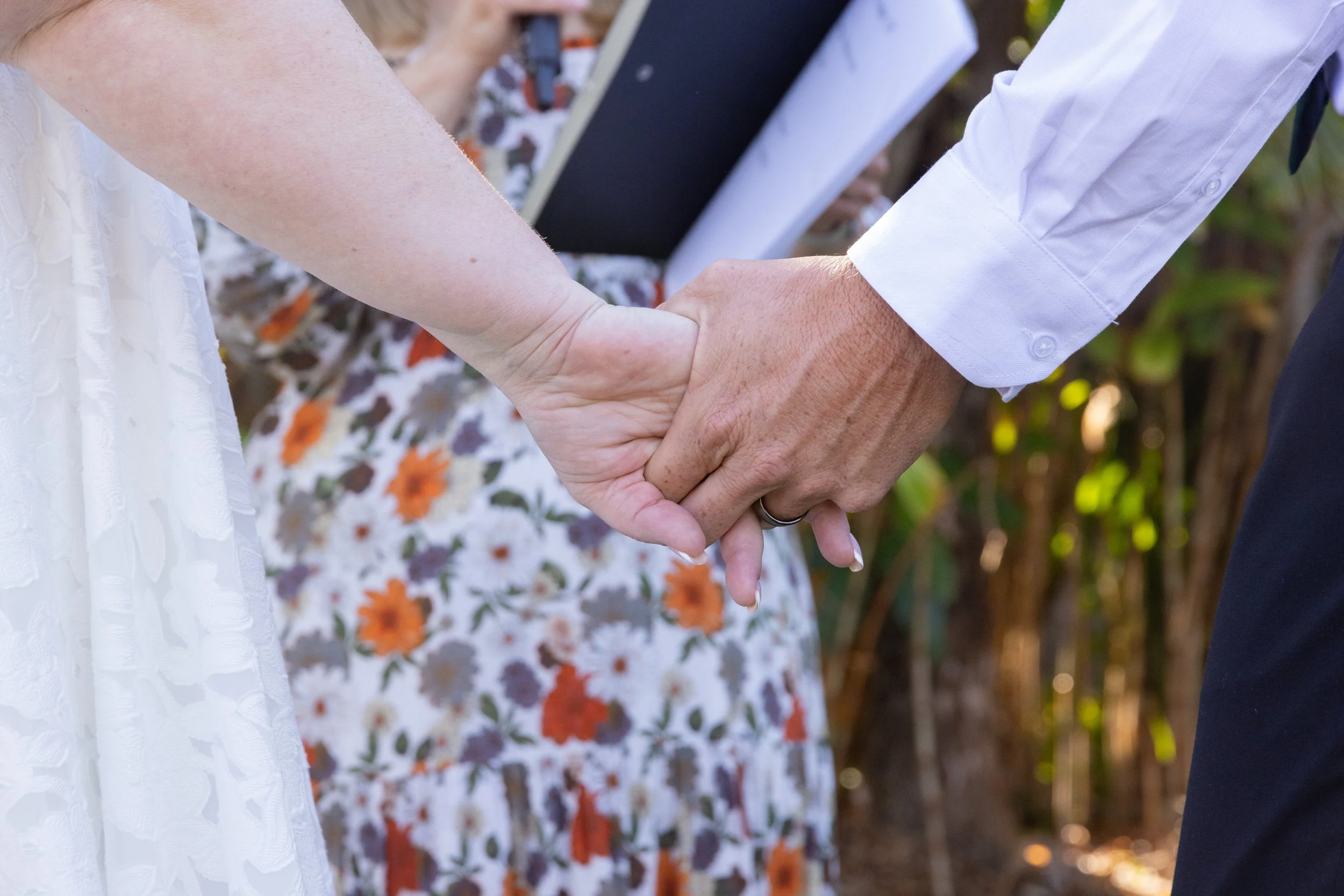 Close-up of a couple holding hands during a wedding ceremony, with a person in a floral dress and an officiant in the background.