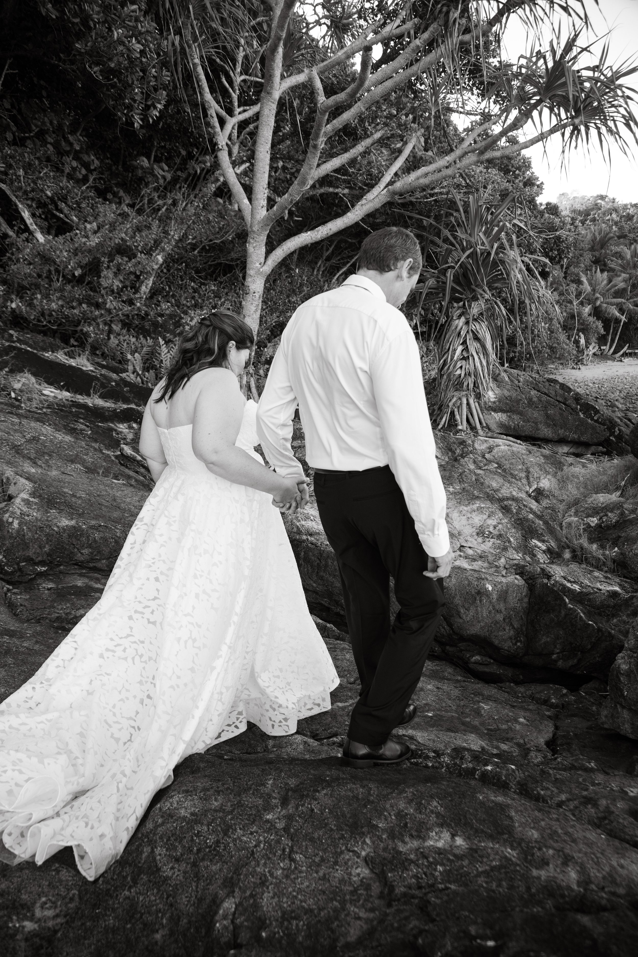 A black-and-white photo of a couple in wedding attire, holding hands and walking on rocks near a beach with trees in the background.