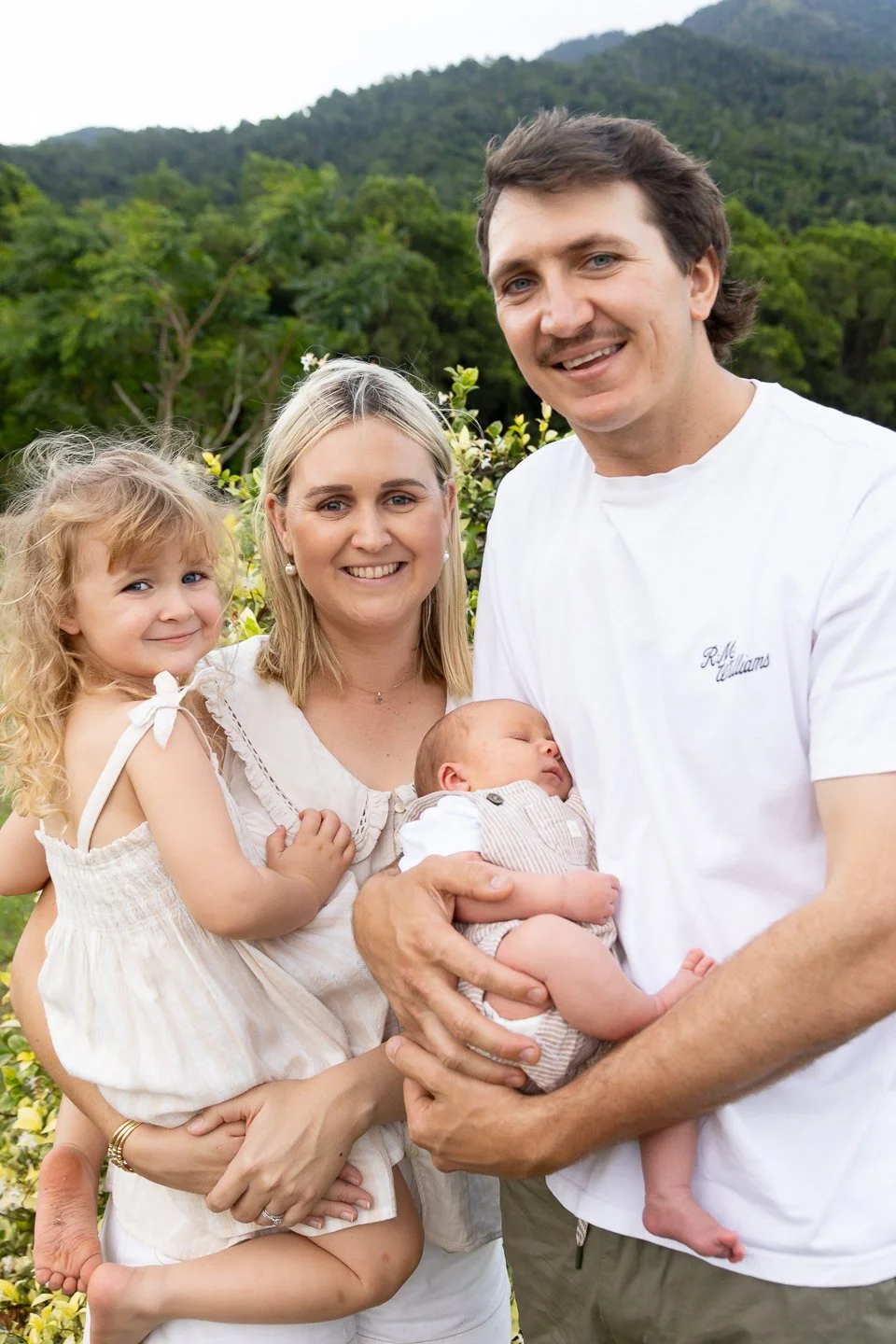 A family of four outdoors in a lush green landscape, including a man holding a sleeping baby, a woman, and a young girl smiling.
