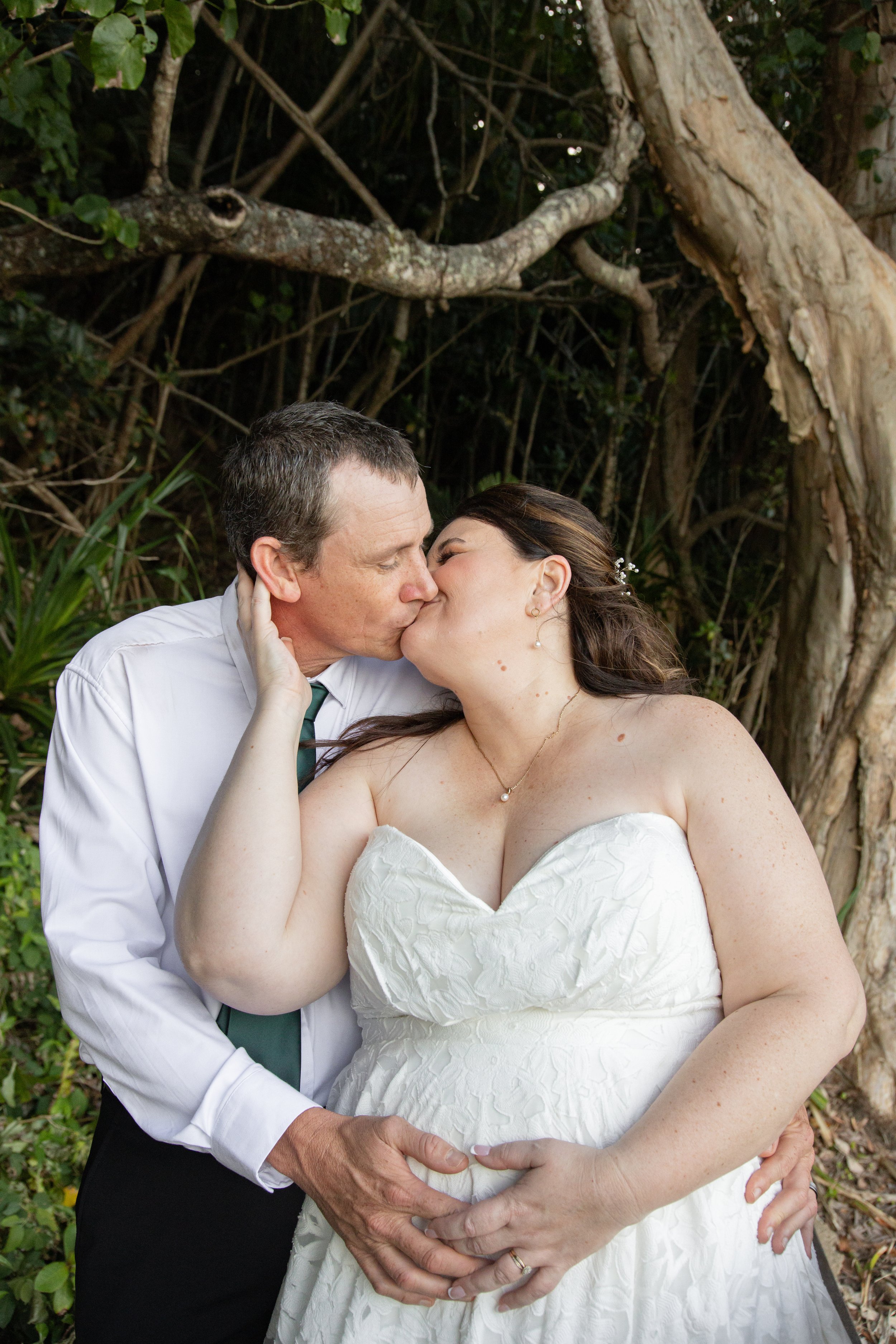 A newlywed couple sharing a kiss outdoors, with greenery and twisted tree branches in the background.