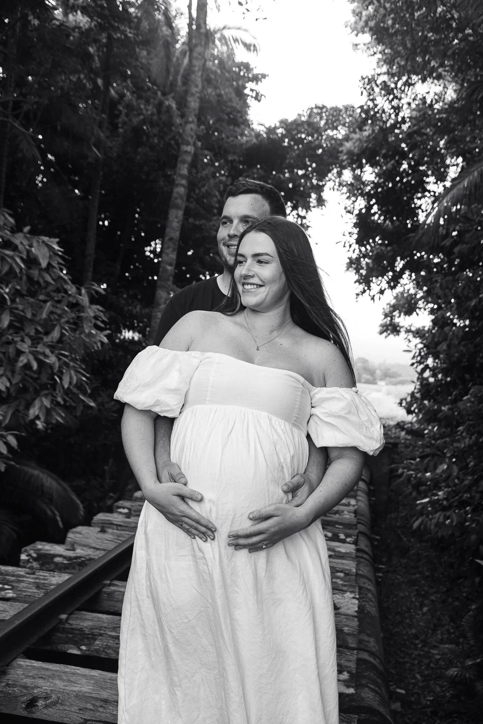 A pregnant woman in a white off-shoulder dress and a smiling man stand on a wooden bridge outdoors, surrounded by trees and foliage, with both hands resting on her belly.