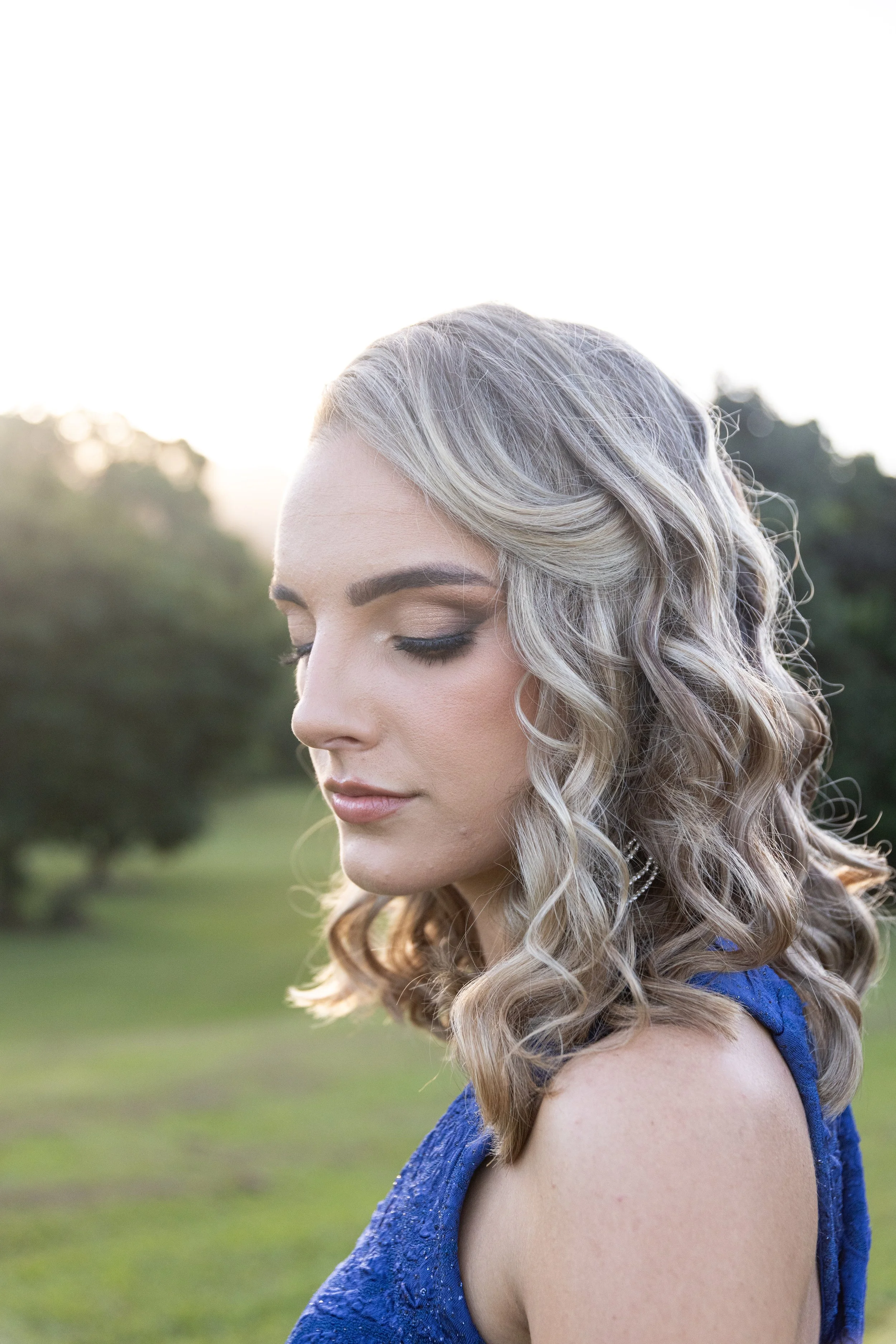 Close-up of a woman with blonde, curly hair and makeup, wearing a blue dress, outdoors in a grassy area during sunset.
