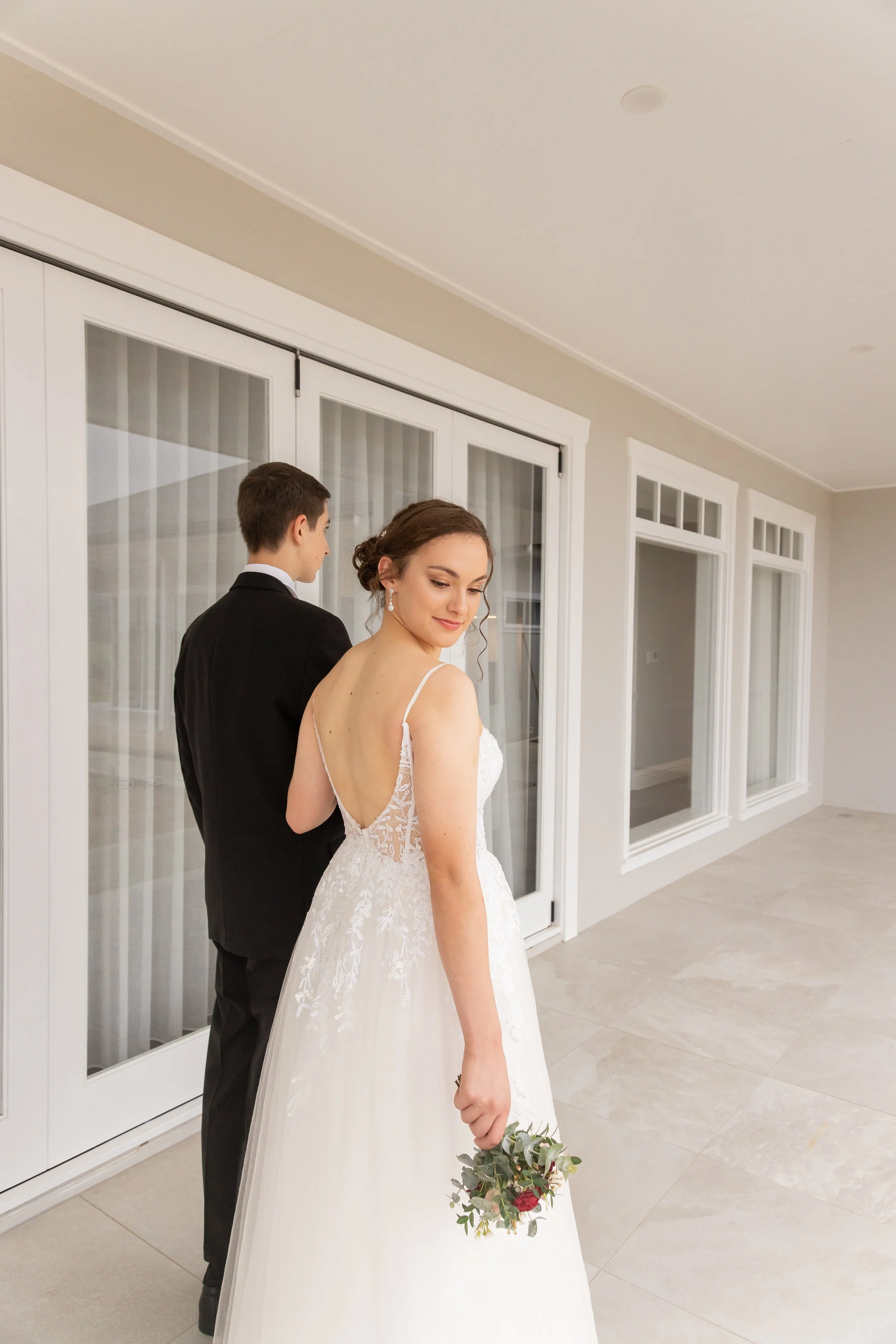 A bride in a wedding dress holding a bouquet, standing next to a groom in a tuxedo, on a porch or patio with sliding glass doors in the background.