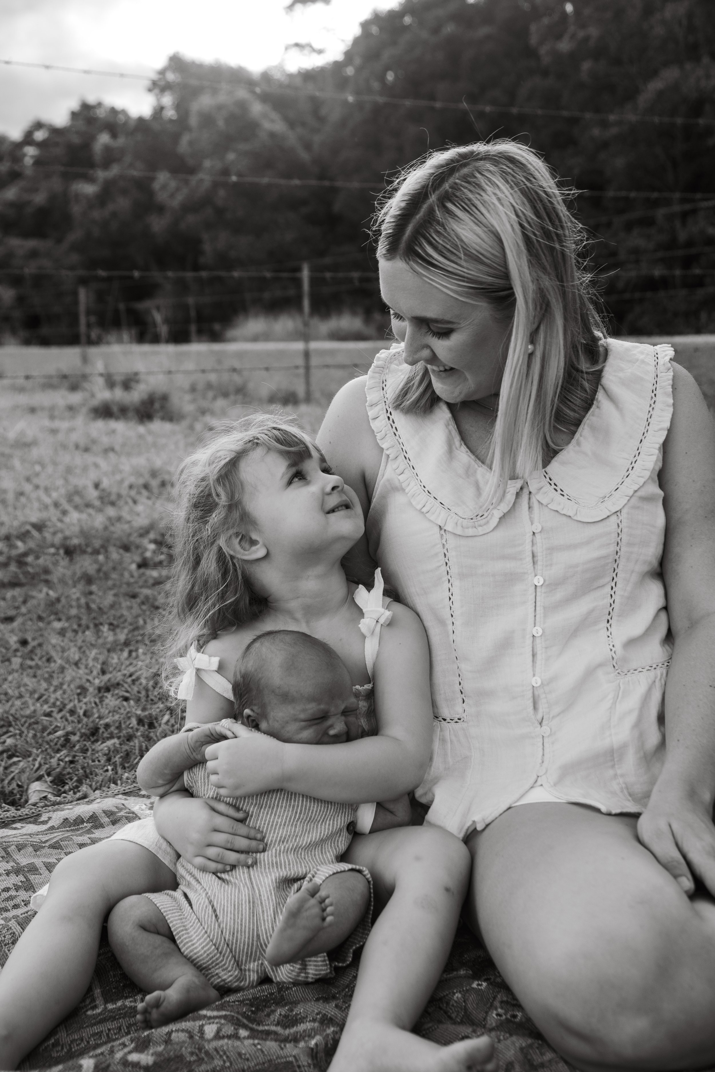 A woman smiling at two young girls, one older girl and a newborn baby, sitting on grass outdoors.