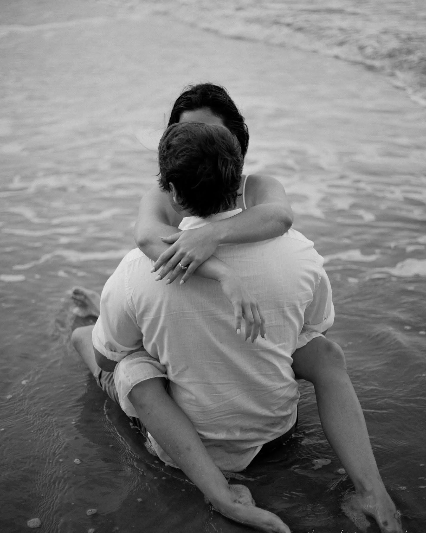 A couple embracing in the water at the beach, with the woman sitting on the man's lap, both with their arms around each other.