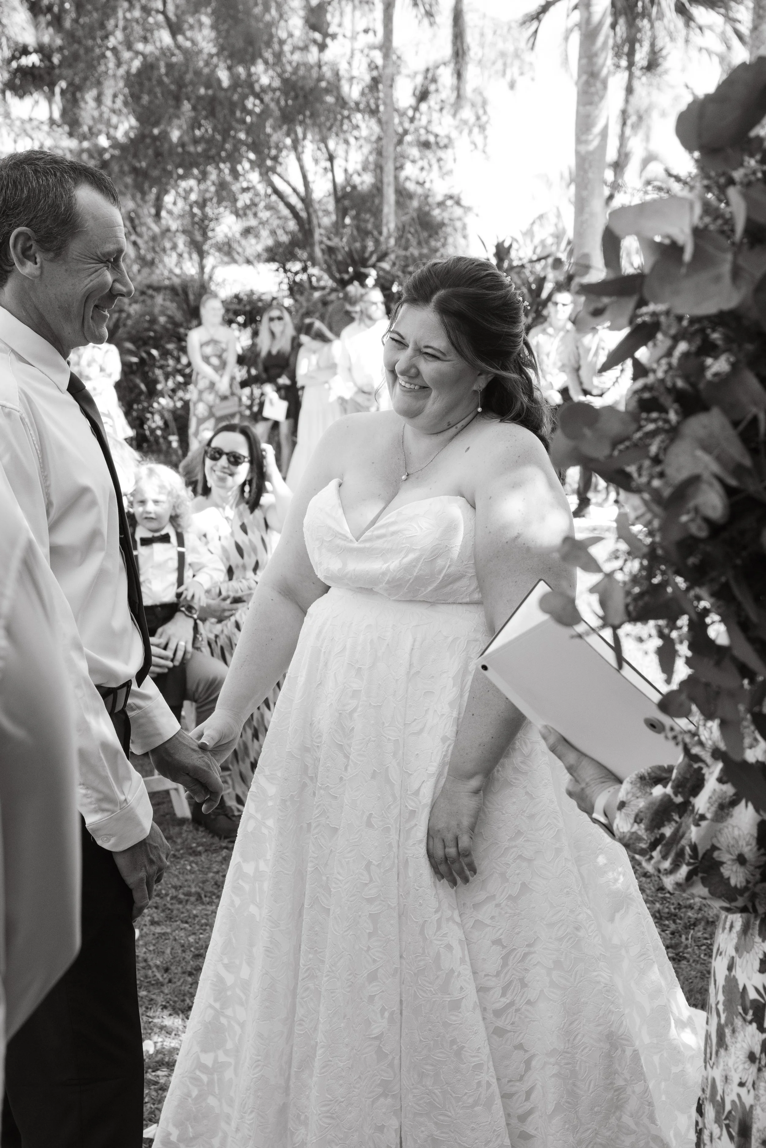 A bride and groom holding hands during a wedding ceremony outdoors, smiling at each other, with wedding guests in the background.
