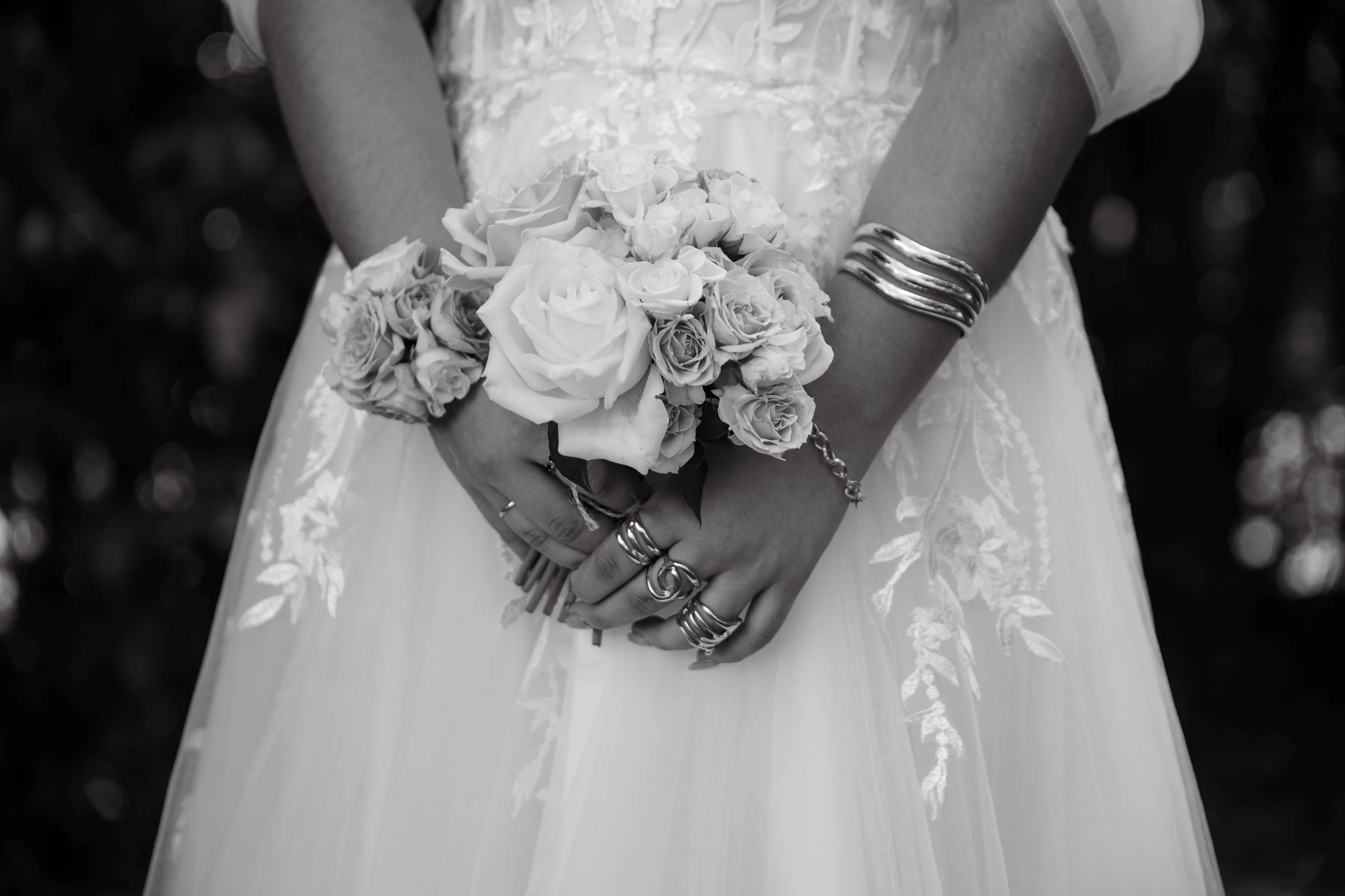 Close-up of a bride holding a bouquet of roses, wearing multiple silver bangles and rings, in a wedding dress with floral embroidery, in black and white.