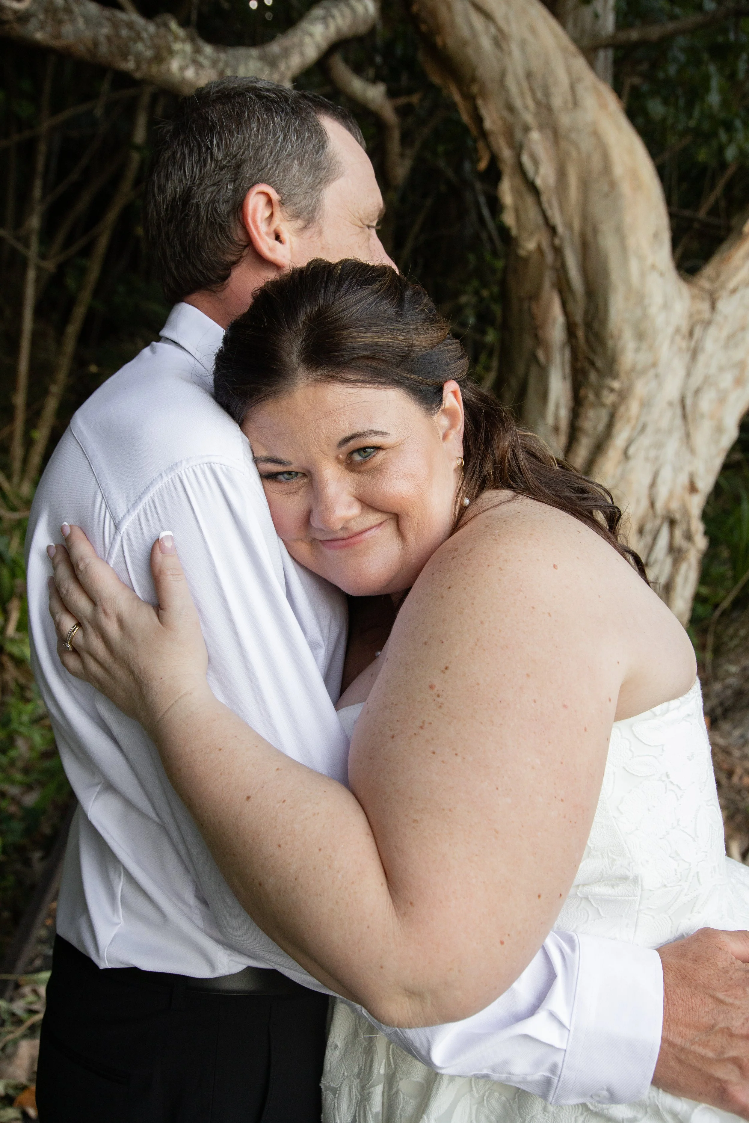 A woman with light skin and dark hair hugging a man with light skin and short dark hair near a tree, smiling softly.