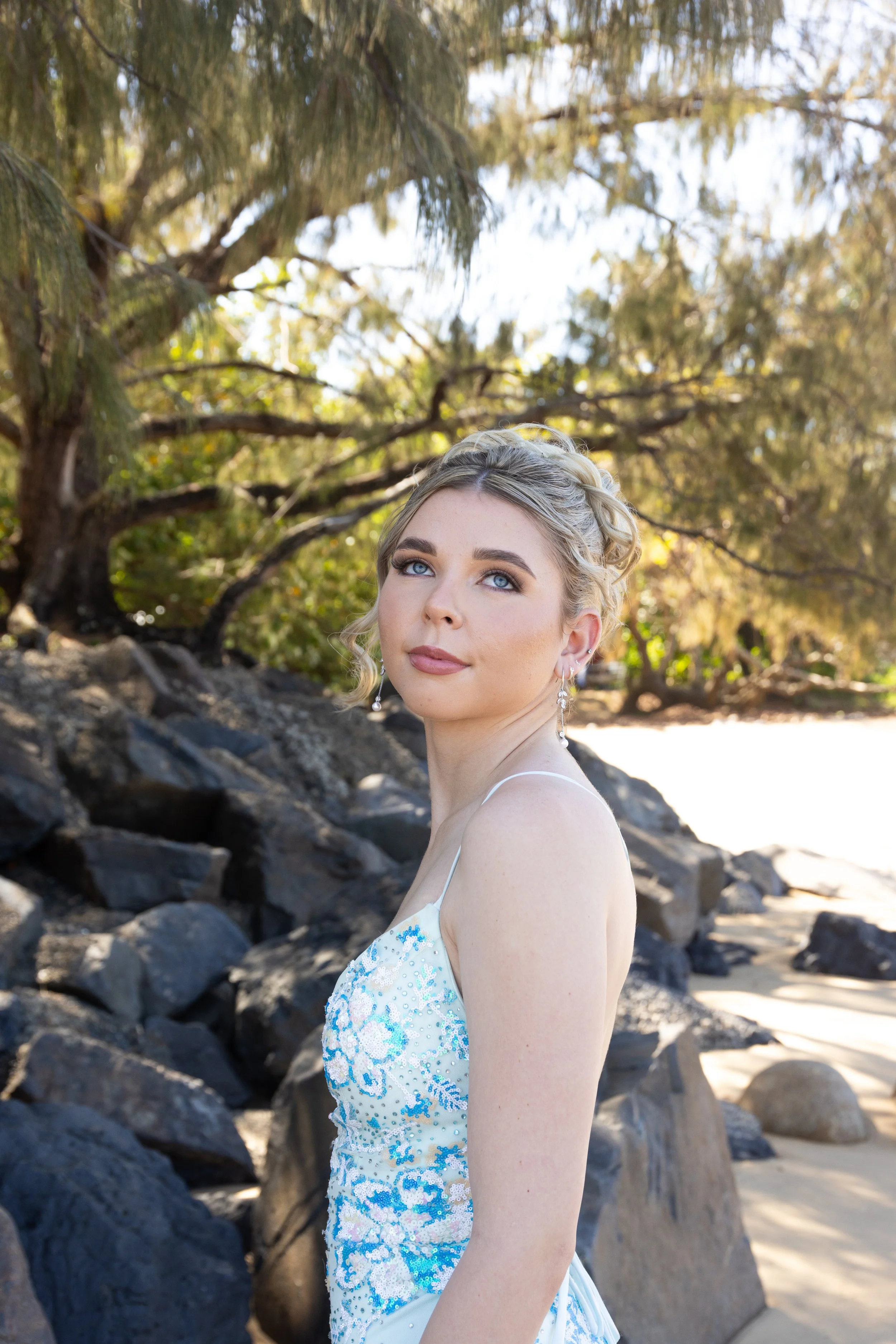 A woman in a white dress with blue accents standing outdoors near rocks with trees and sunlight in the background.