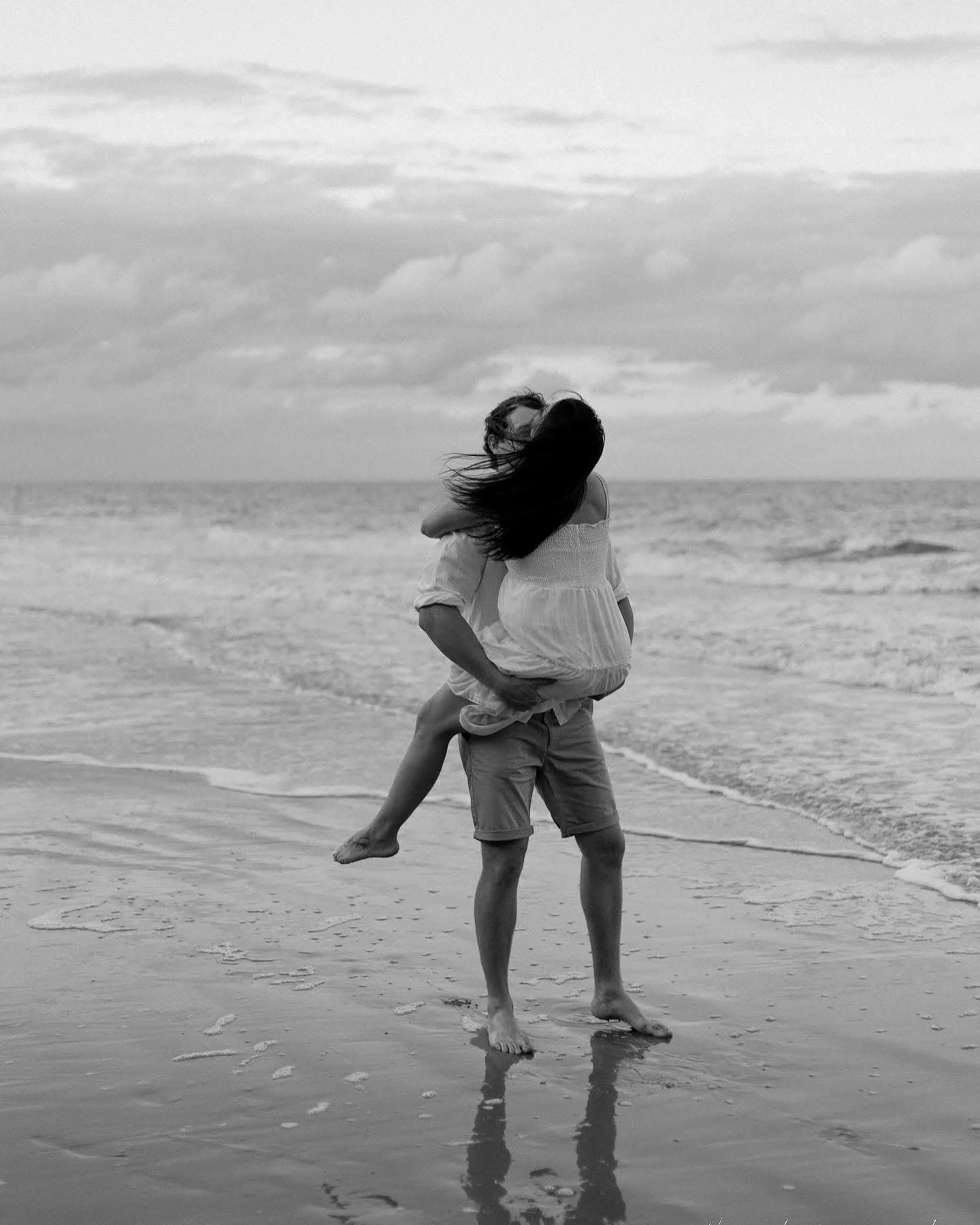 A black-and-white photo of a man lifting a woman in a dress on the beach, with the ocean and cloudy sky in the background.