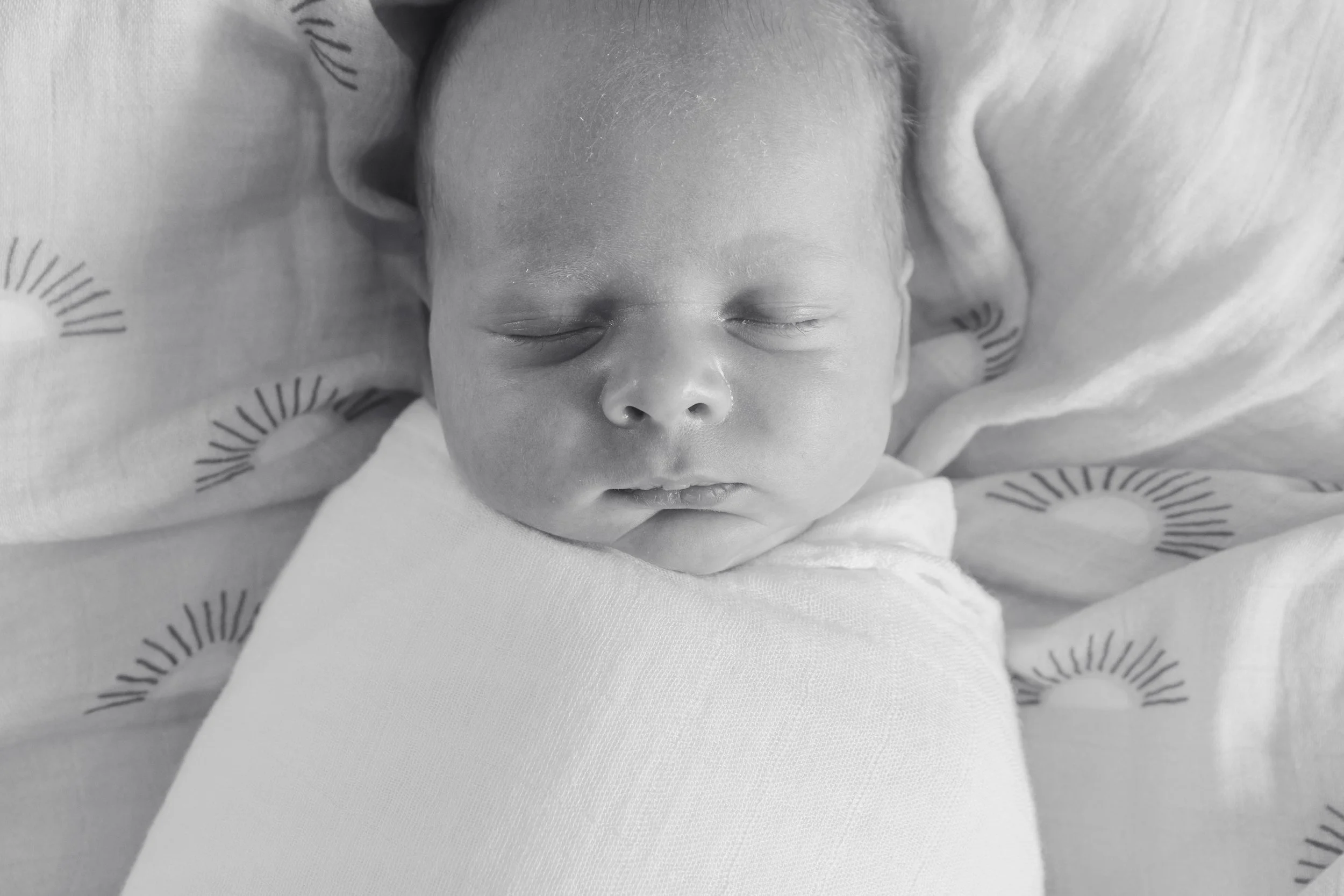 Black and white photo of a sleeping newborn baby wrapped in a white blanket, lying on a patterned sheet.