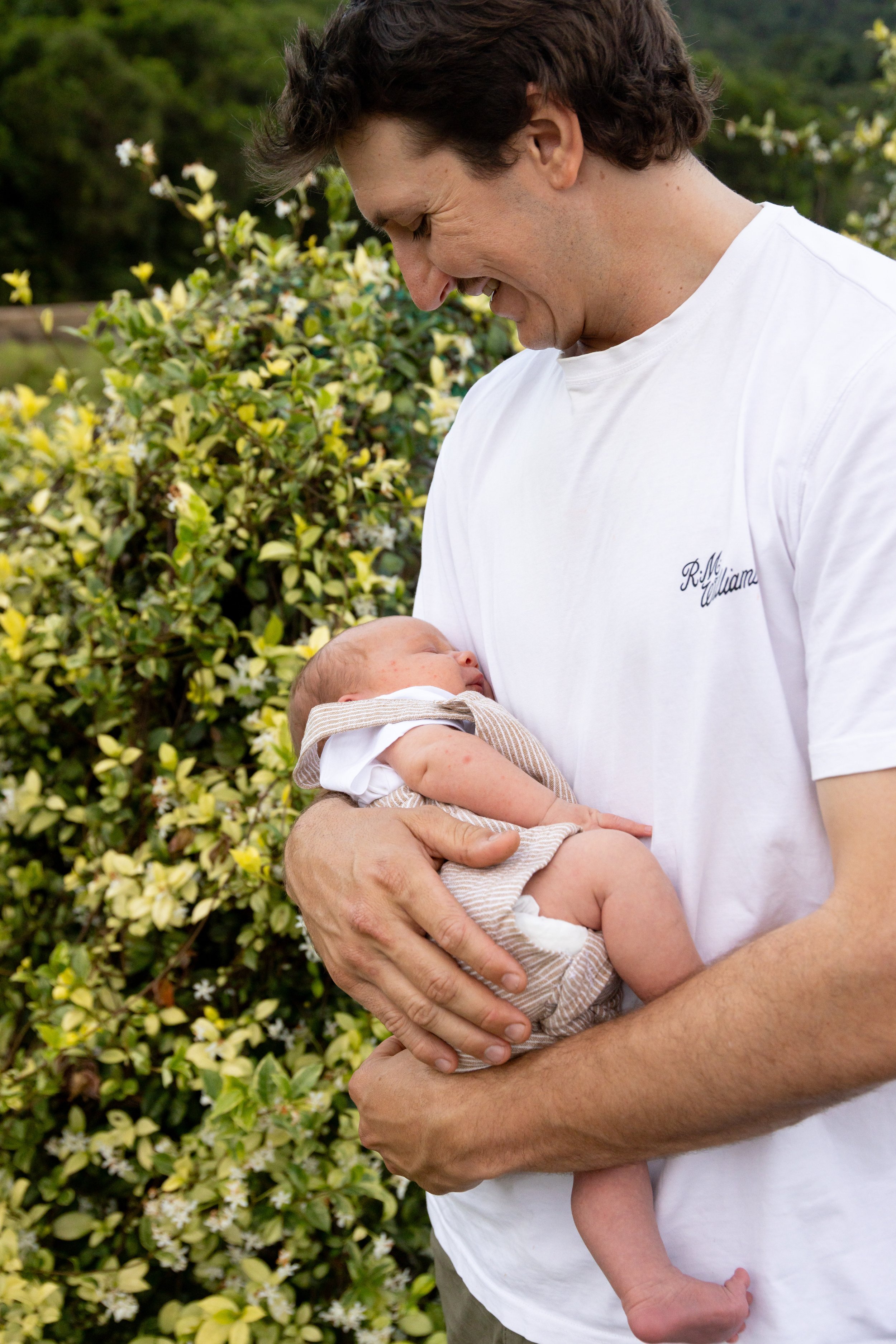 A man holding a newborn baby outdoors, with green bushes and yellow flowers in the background.