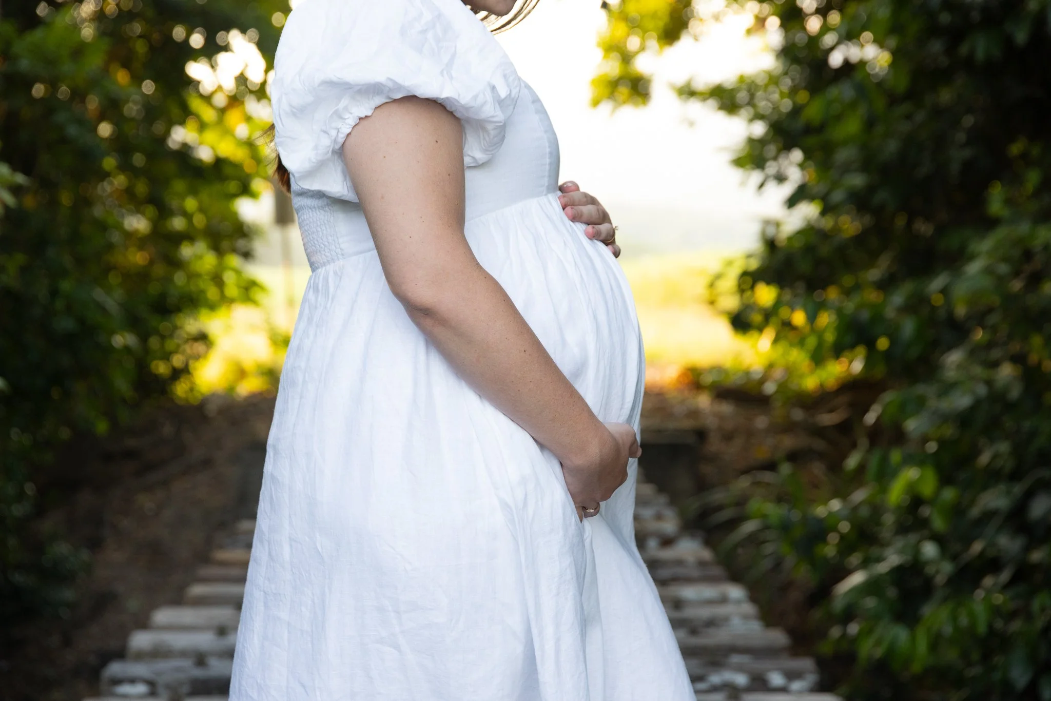A pregnant woman in a white dress standing on a wooden bridge outdoors, holding her belly with one hand, surrounded by greenery.
