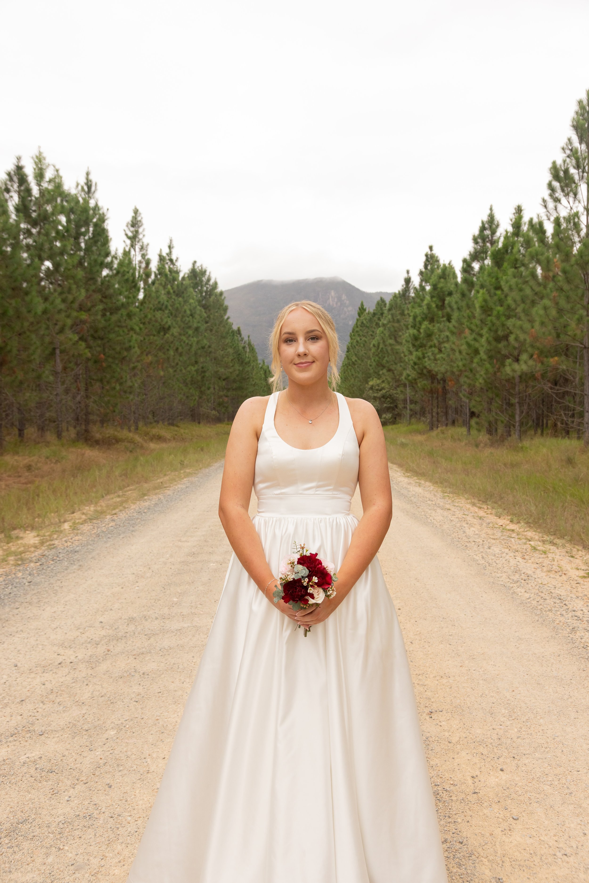 A woman in a white wedding dress holding a bouquet of red and white flowers, standing on a dirt road surrounded by green pine trees, with mountains in the background.