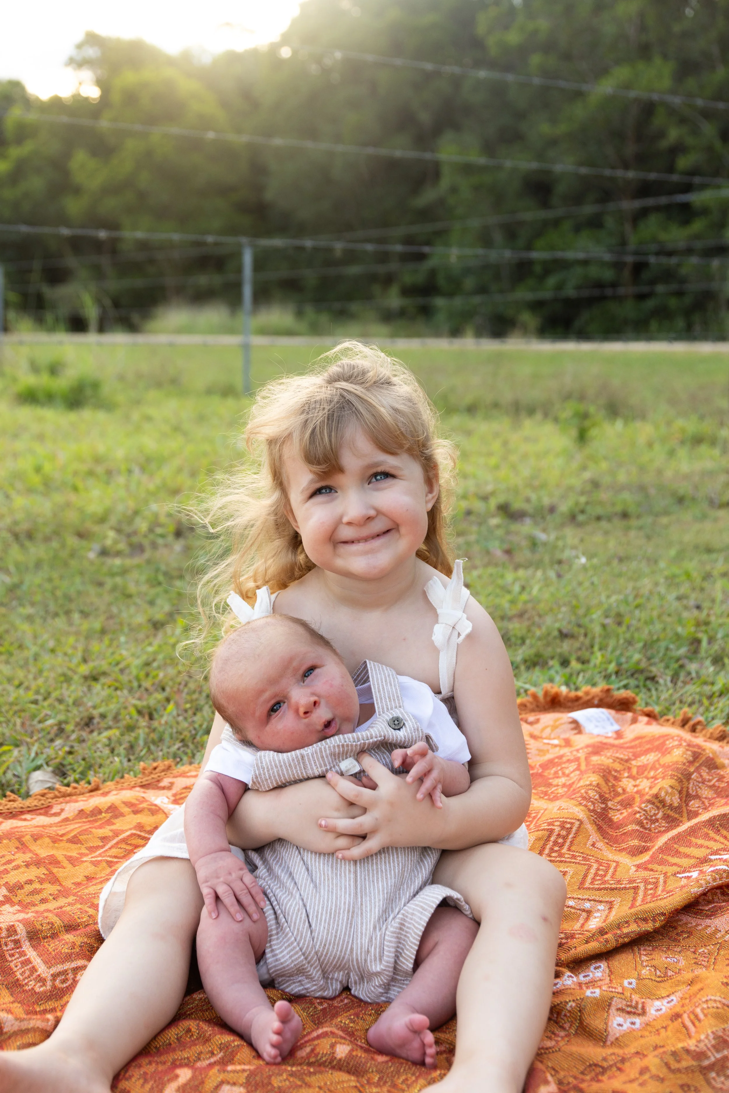 A young girl with blonde hair smiling while holding a newborn baby on her lap, sitting on an orange patterned blanket outdoors on a grassy field during late afternoon.