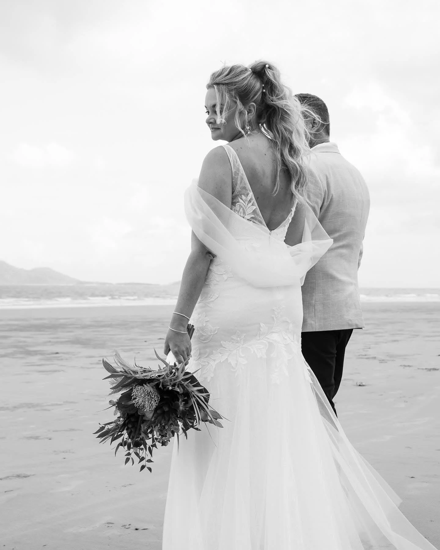 A bride in a wedding dress holding a bouquet standing on a beach with a groom in a light-colored suit facing away, under a cloudy sky.