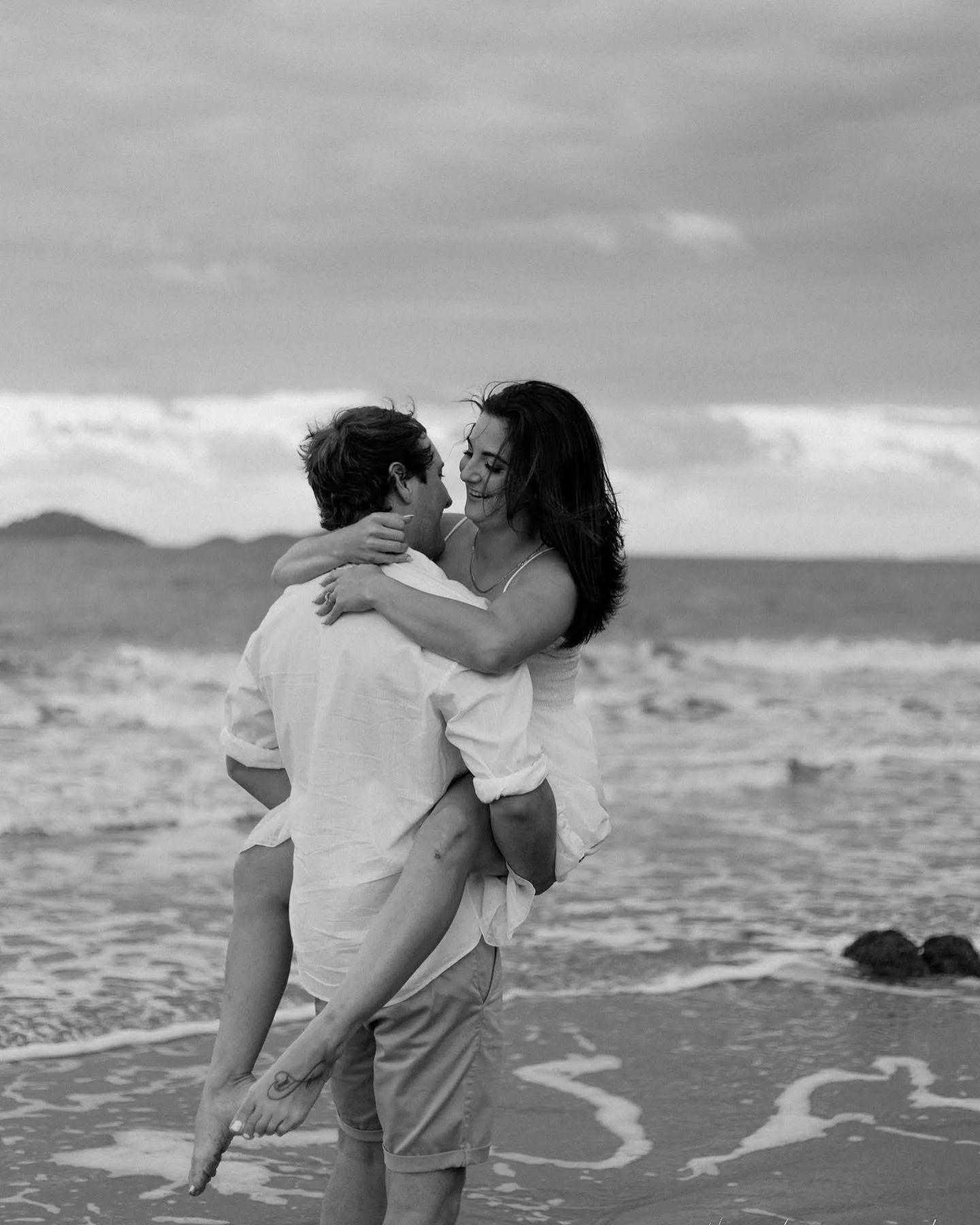 A couple is at the beach, with the man holding the woman in his arms, both smiling and looking at each other.
