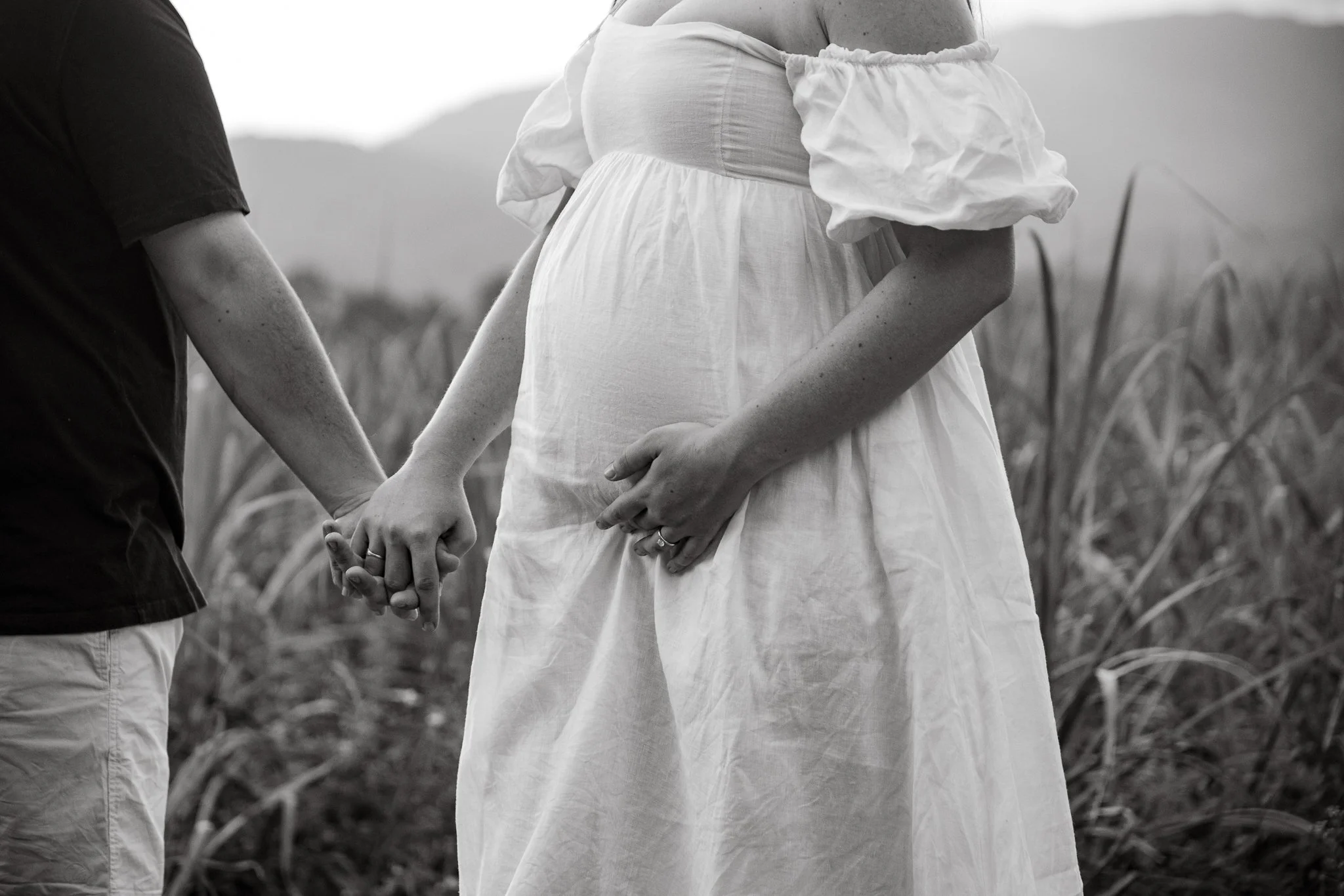 A pregnant woman and another person holding hands in a field, with the woman wearing a white dress and cradling her belly.
