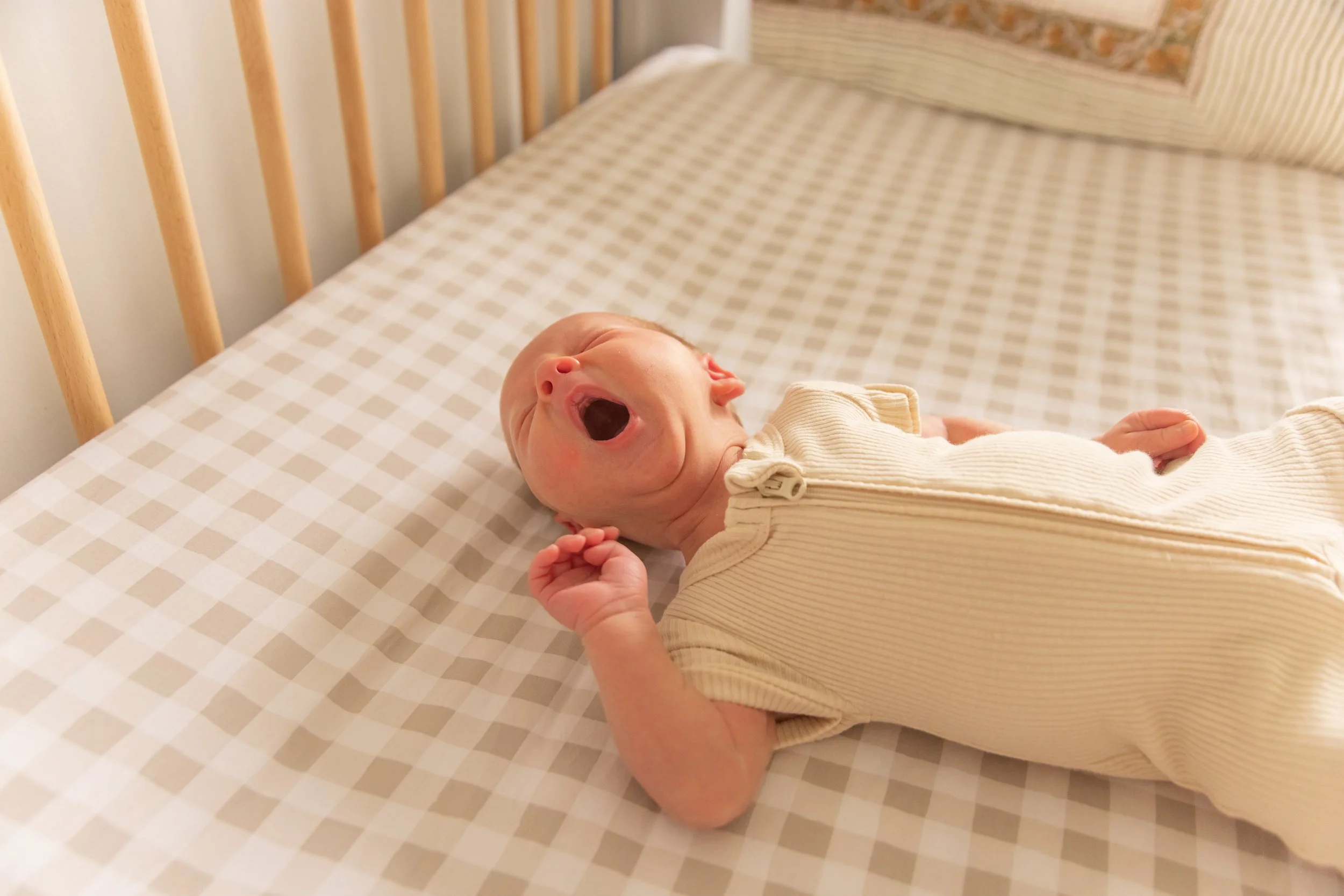 A yawning baby lying on a checkered bed with a wooden headboard and a patterned pillow in the background.