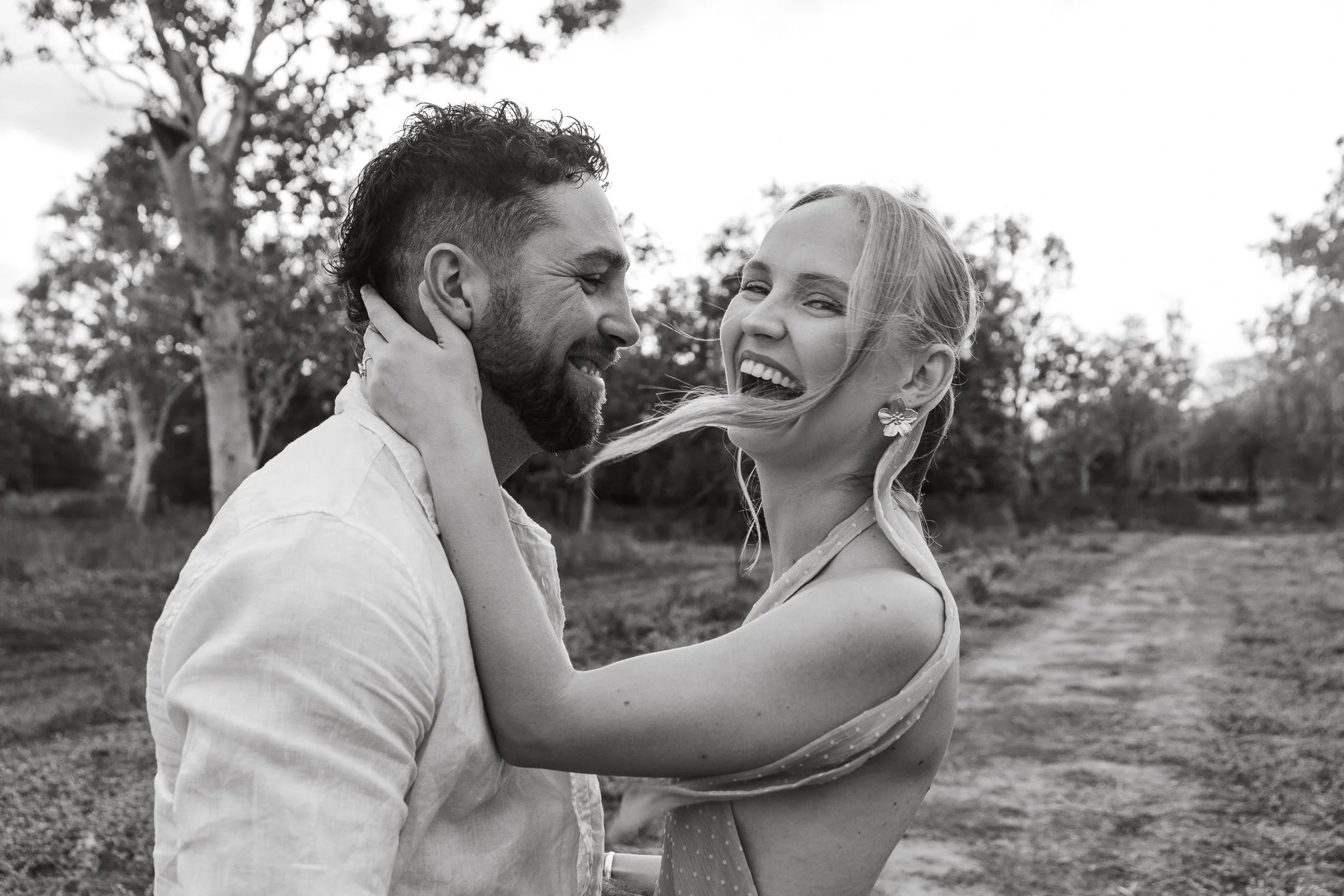 A black and white photo of a couple laughing outdoors, with trees in the background. The woman is holding the man's face with one hand and wearing a sleeveless dress, earrings, and has her hair flowing. They are looking at each other and smiling.