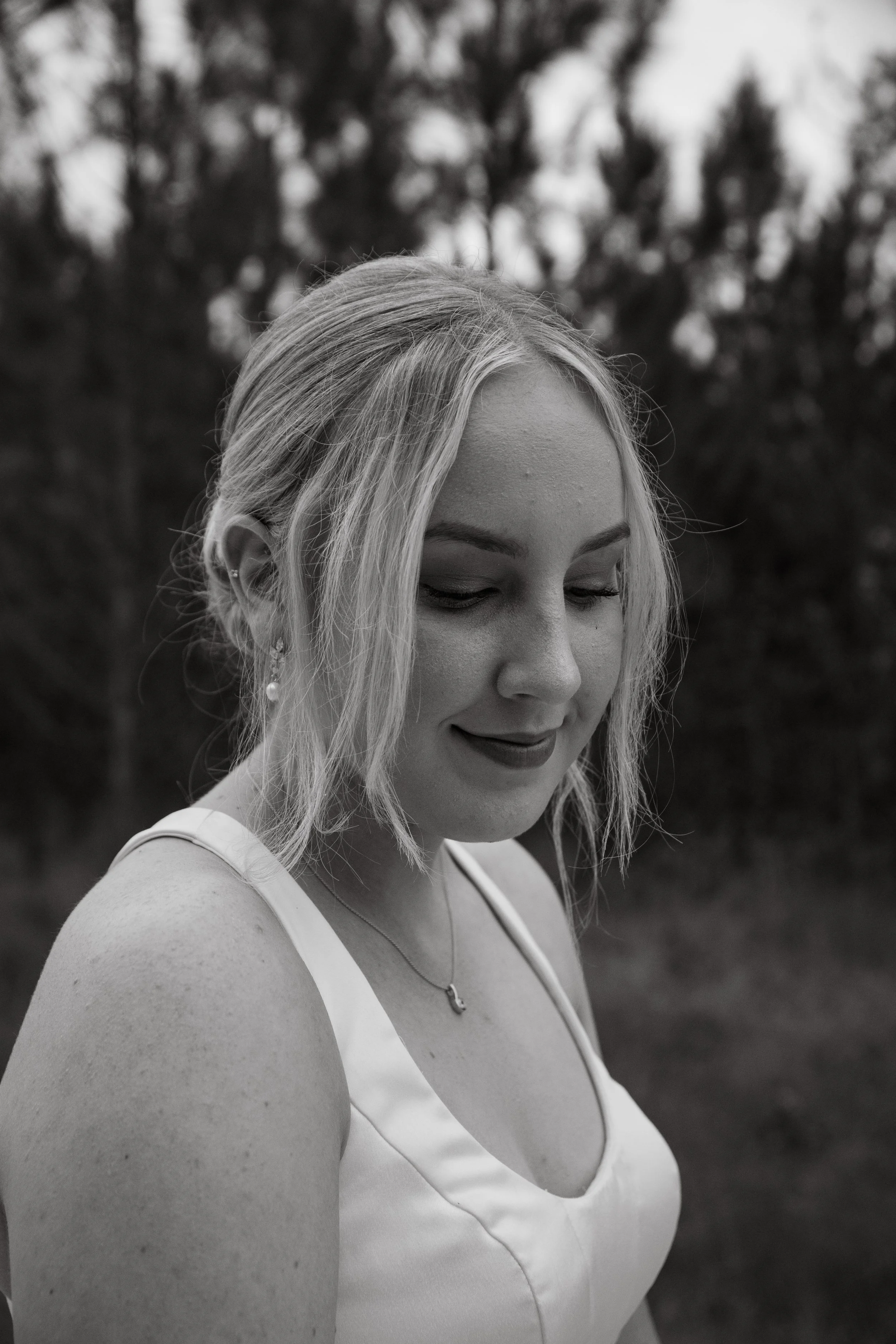 A young woman with blonde hair, wearing a white dress, earrings, and a necklace, looking down outdoors.