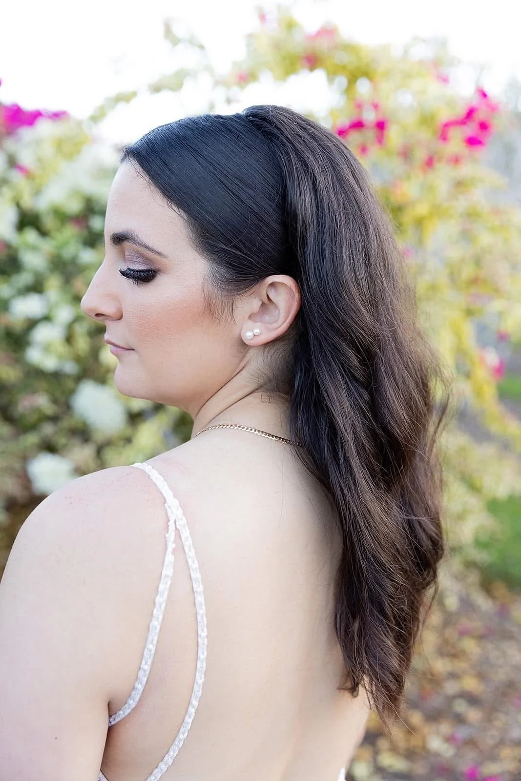 Side profile of a woman with dark wavy hair, wearing pearl earrings and a white lace strap dress, standing outdoors with pink and white flowers in the background.