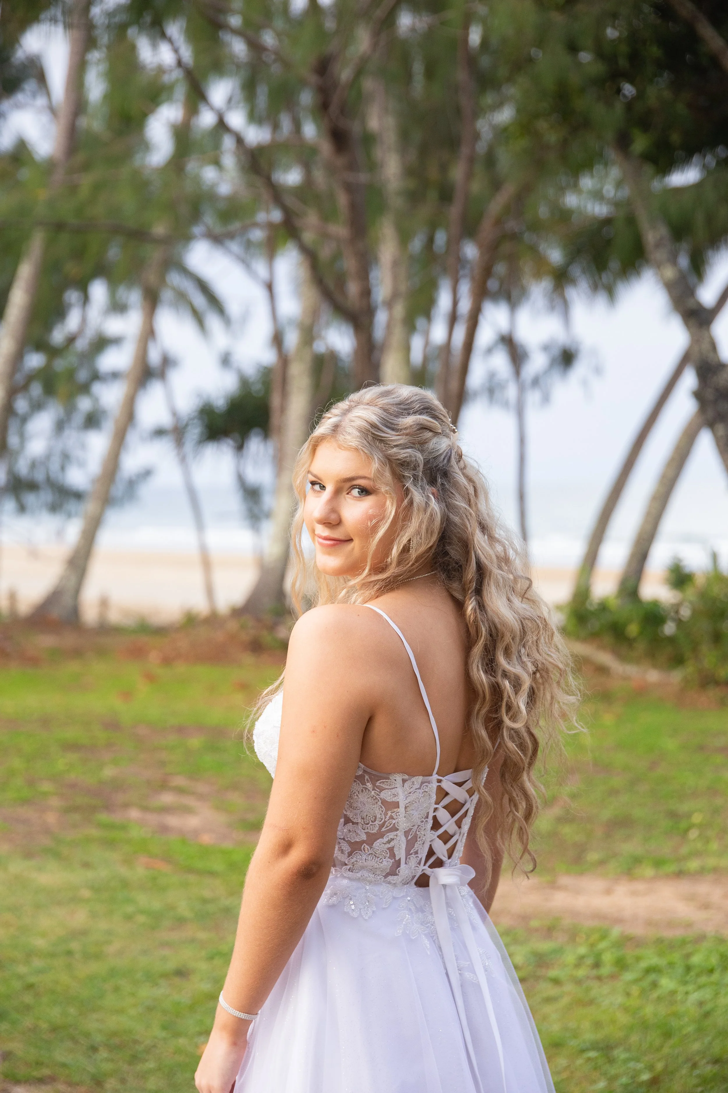 A young woman with long, curly blonde hair in a white dress, standing outdoors near trees and a beach.