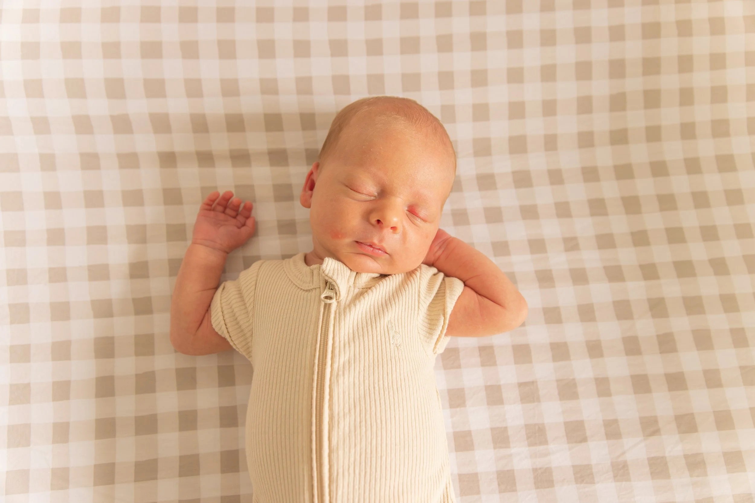 A sleeping newborn baby lying on a beige checkered surface, wearing a cream-colored zip-up onesie.