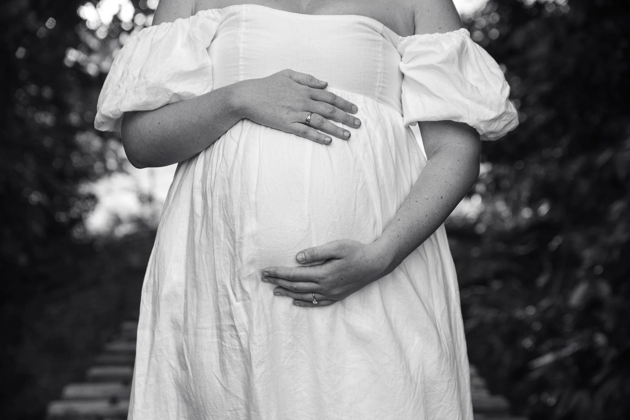 Black and white photo of pregnant woman in a white dress, with her hands on her belly, outdoors with blurred trees in background.