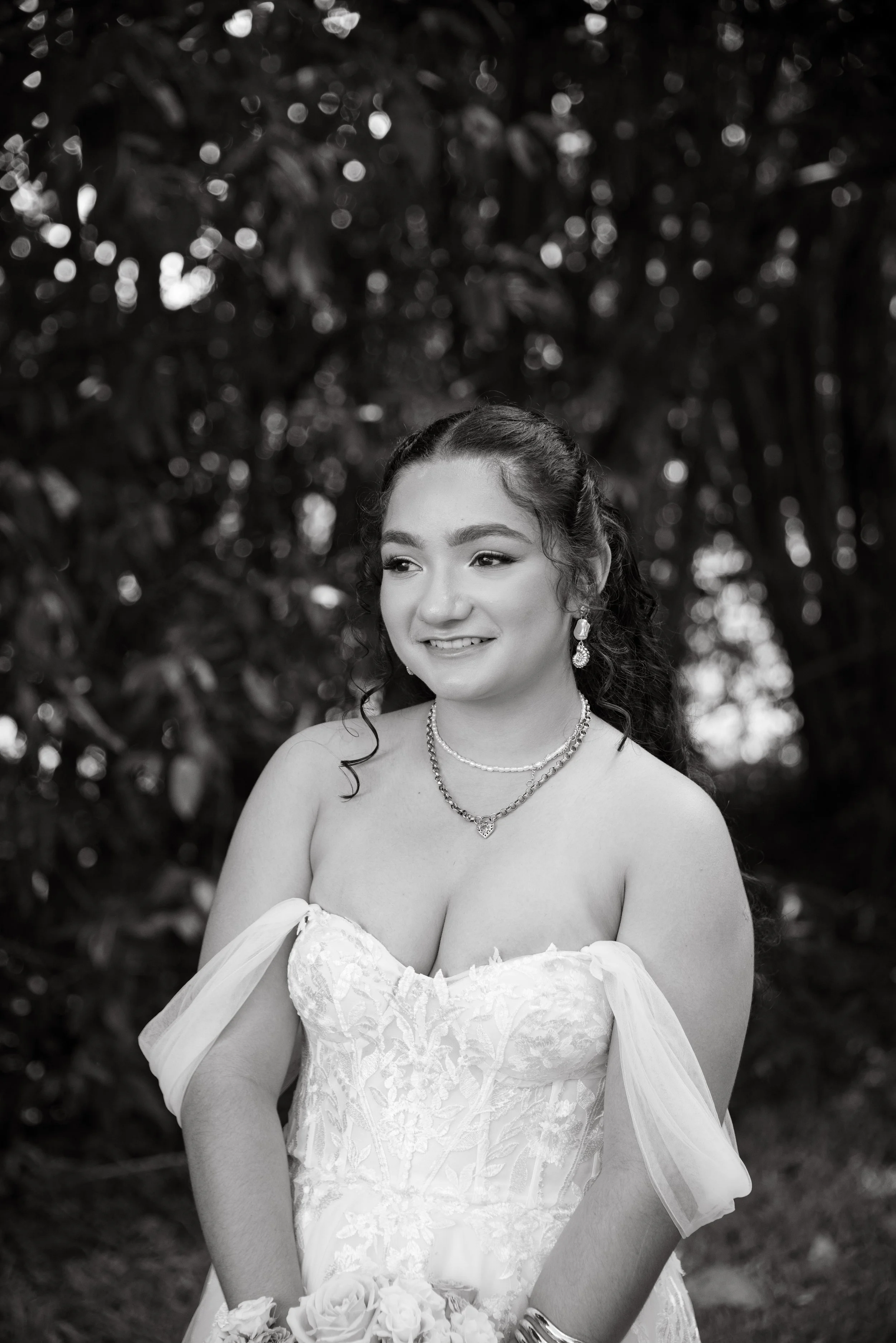 Black and white portrait of a young woman in a wedding dress outdoors, smiling and holding a bouquet of roses, with a blurred background of trees.