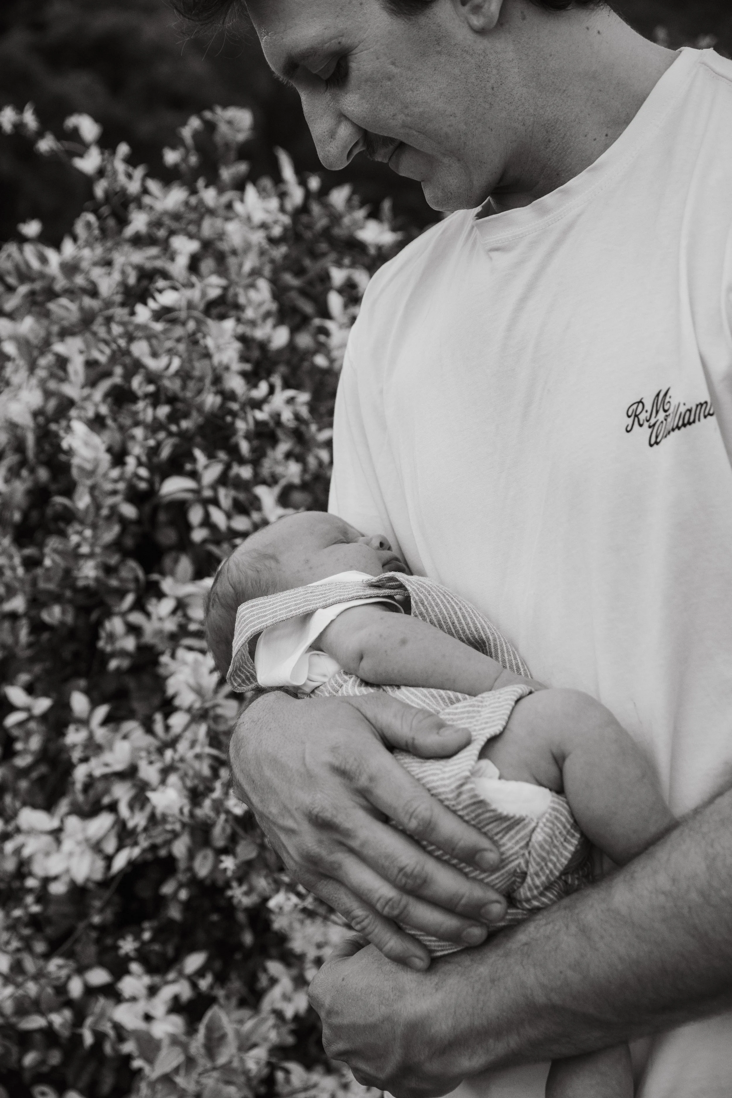 A person holding a newborn baby outdoors, with a background of flowers, in black and white.