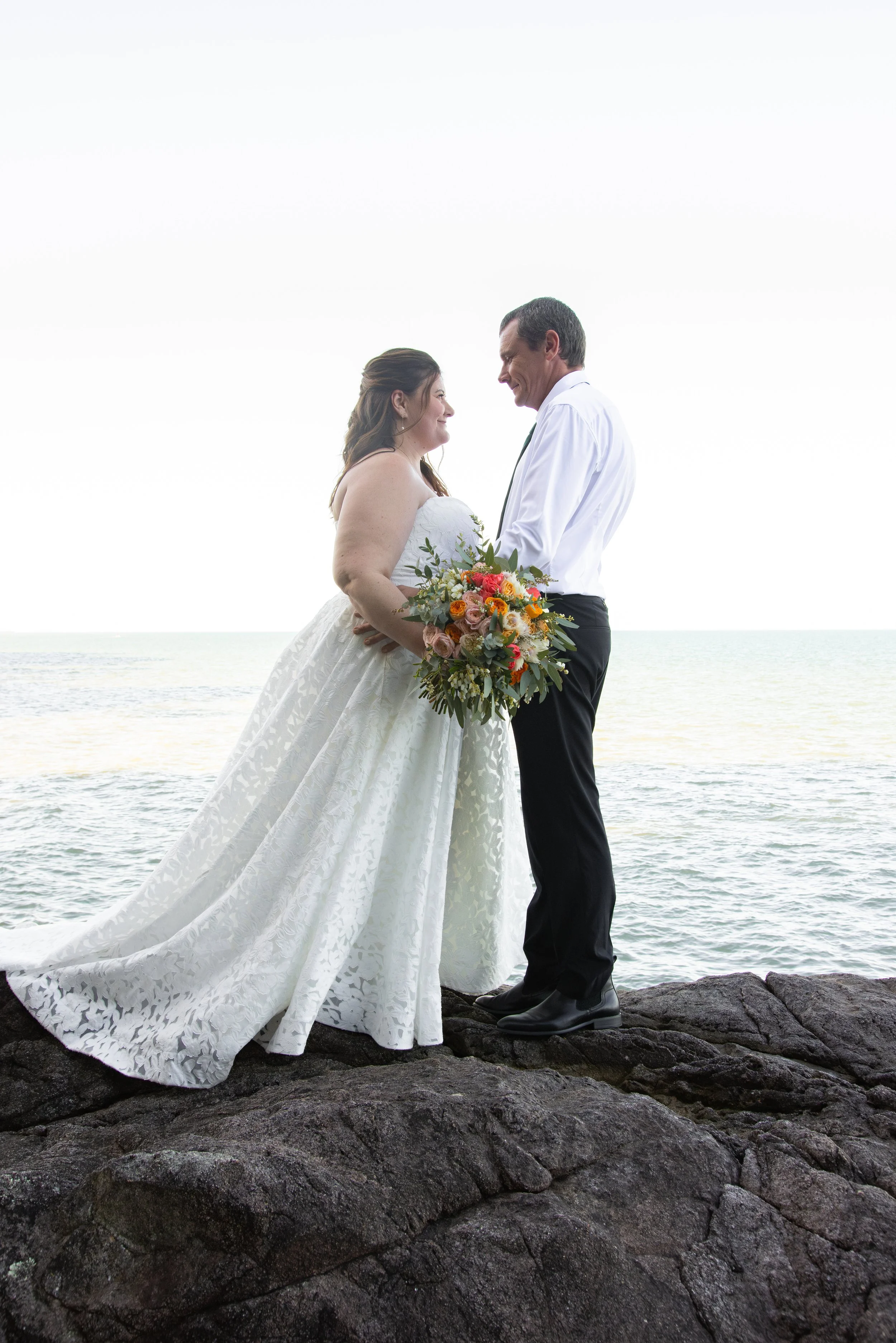 A bride and groom on a rocky shoreline, holding hands and gazing at each other, with an ocean in the background and the sky overcast.
