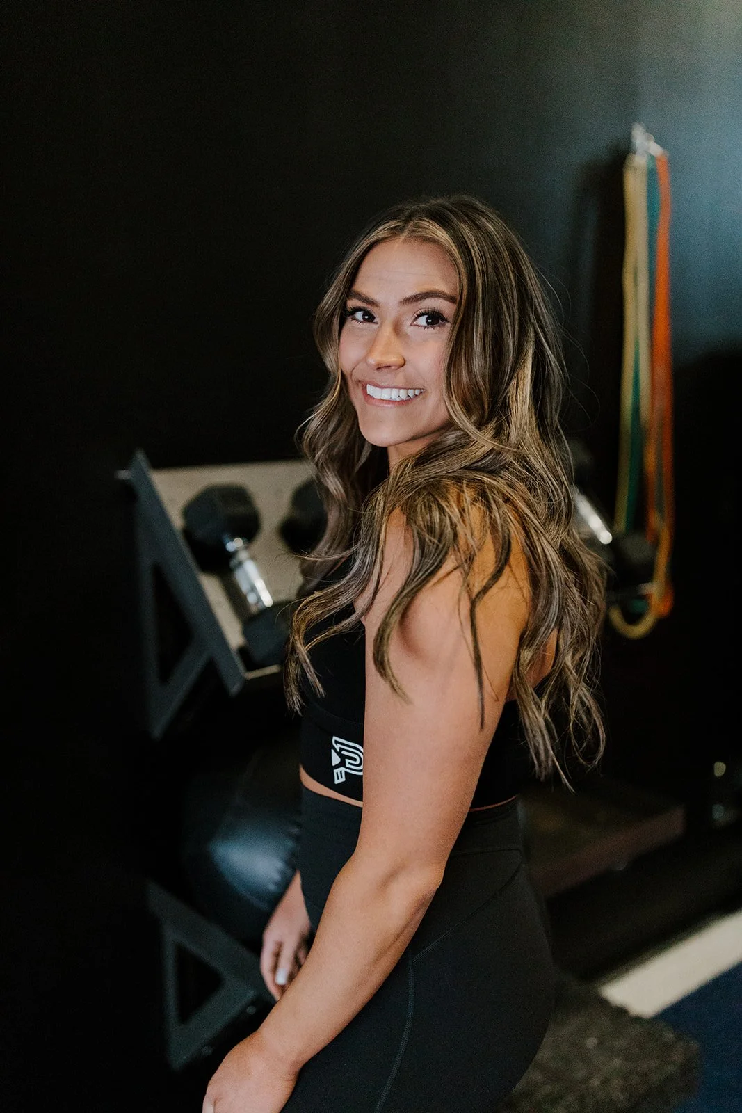 A young woman with long, wavy brown hair smiling at the camera, standing in a fitness or gym setting with dumbbells and resistance bands in the background.